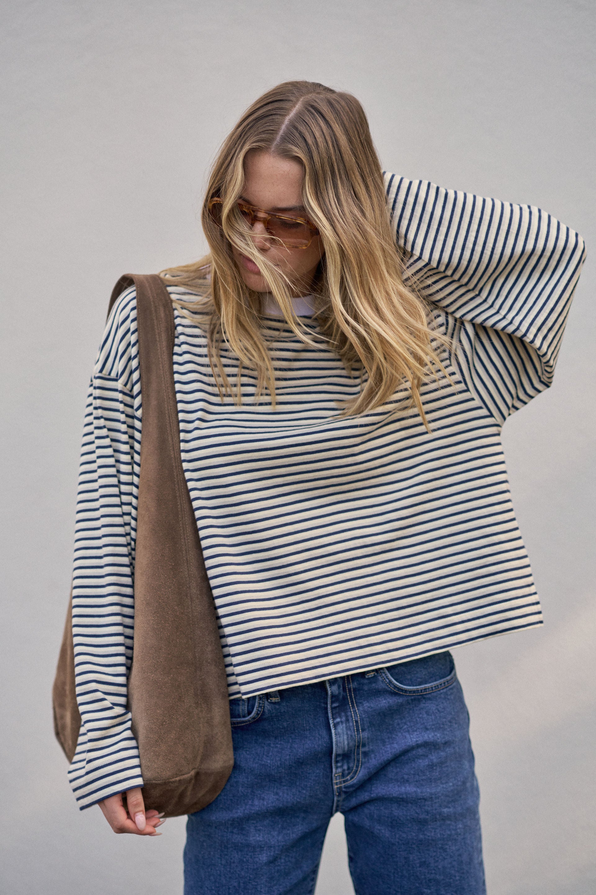 Woman wearing a Ceres Life striped sweater and blue jeans, holding a brown bag against a plain background