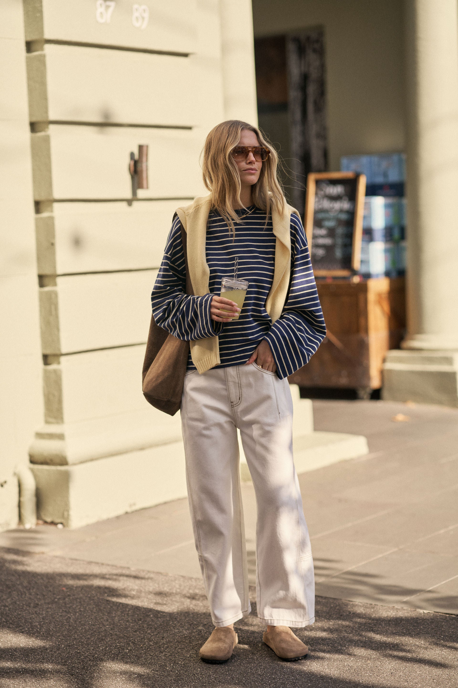 Image of Woman in Ceres Life striped sweater and white pants standing on a sidewalk holding a coffee cup.