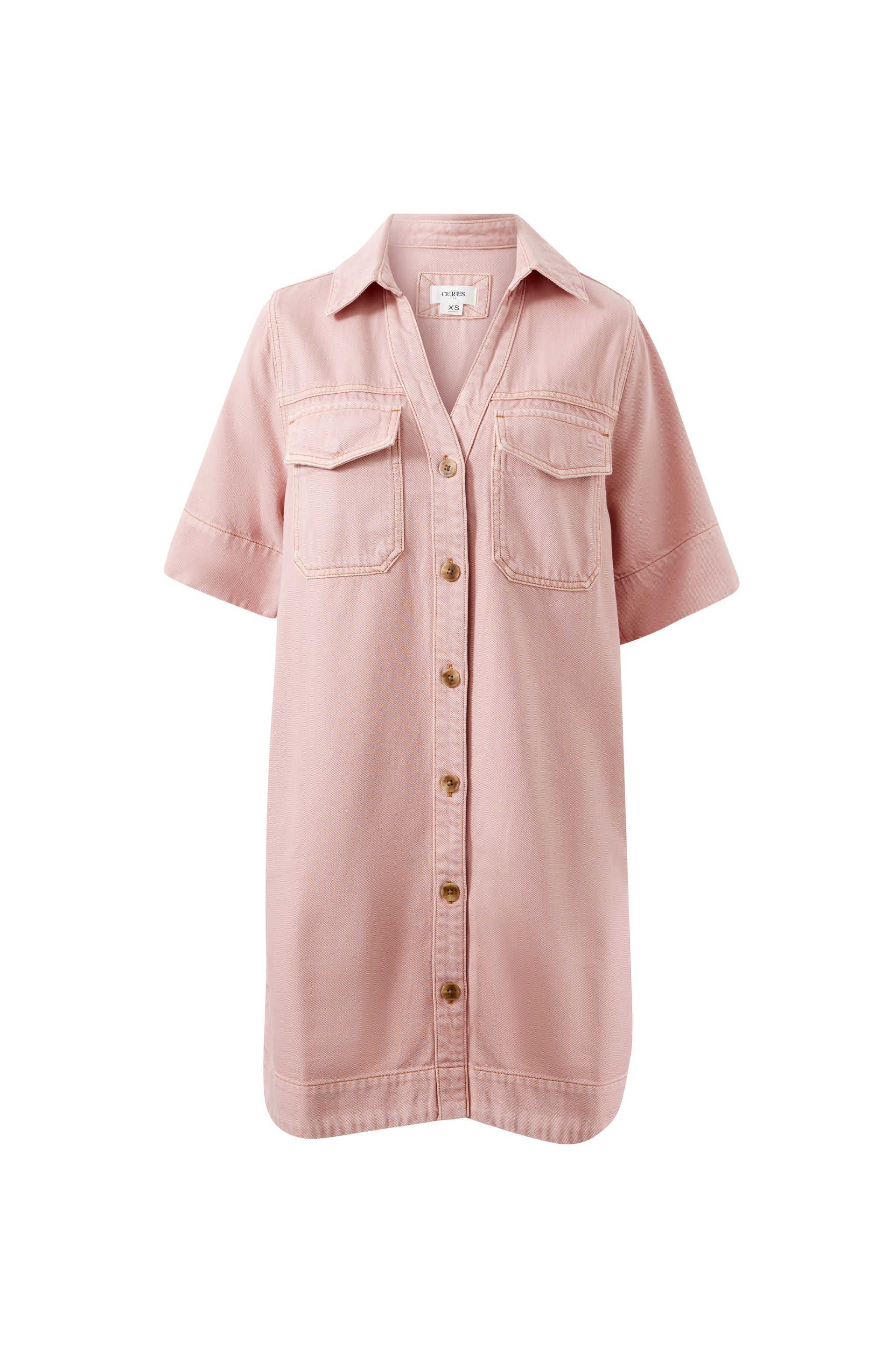Image of Model wears a relaxed pink denim shirt dress featuring a button-through front, short sleeves, chest pockets and a collared neckline, styled with black sandals.