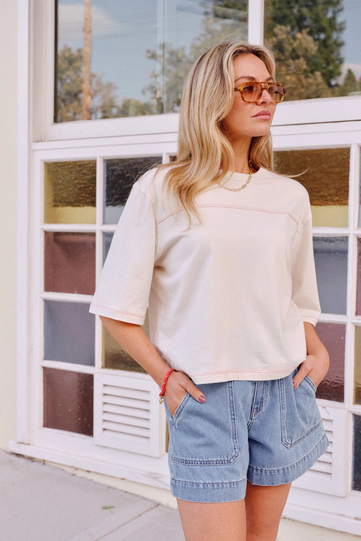 A woman wearing a Ceres Life cream-colored short-sleeve knit top with pinkish-red contrast stitching and light blue denim shorts, standing in front of a white-framed window.
