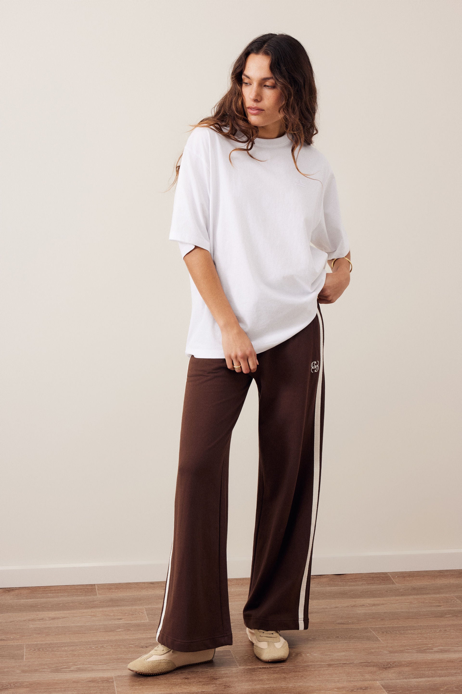 Image of A woman modeling a white oversized short-sleeve t-shirt and Ceres Life dark brown wide-leg sweatpants with white side stripes, standing on a light wood floor against a beige wall.
