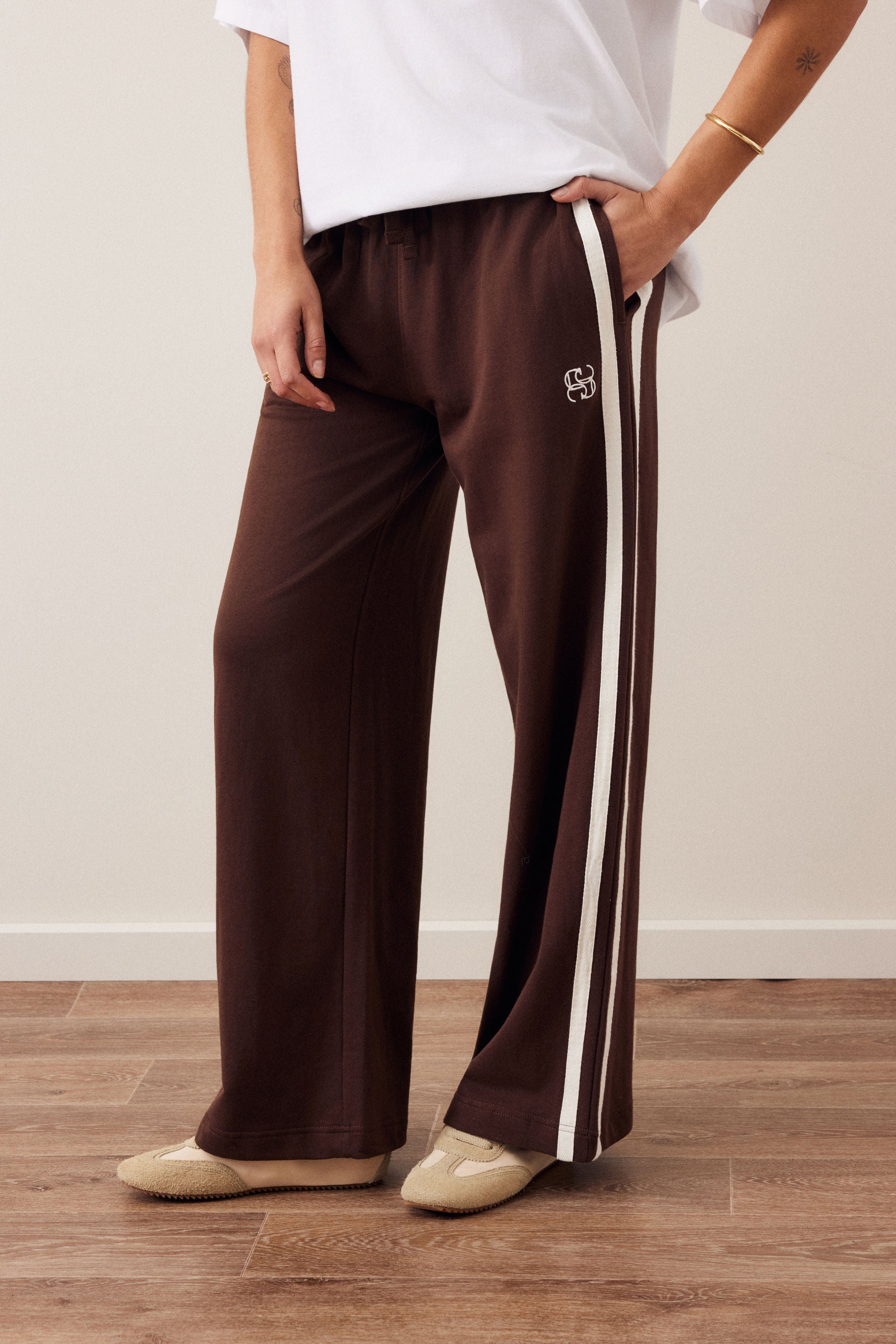 Image of A woman stands outdoors wearing a brown and beige striped knit sweater, Ceres Life brown wide-leg track pants with white side stripes, and brown flat shoes, holding a brown tote bag.