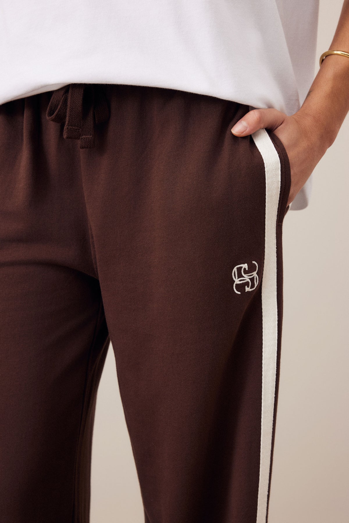 Close-up of a woman wearing Ceres Life dark brown sweatpants with a white side stripe and a small white embroidered monogram logo near the lower leg, paired with a white top against a light beige background.