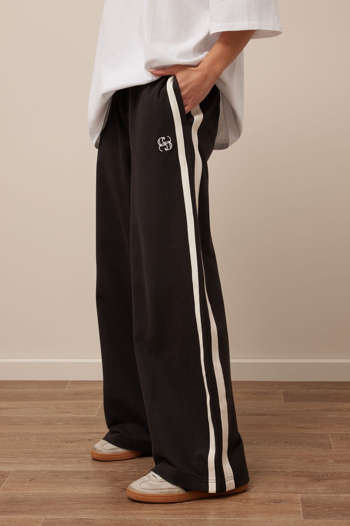 Image of Woman wearing black track pants with white stripes and a logo, standing on a wooden floor.