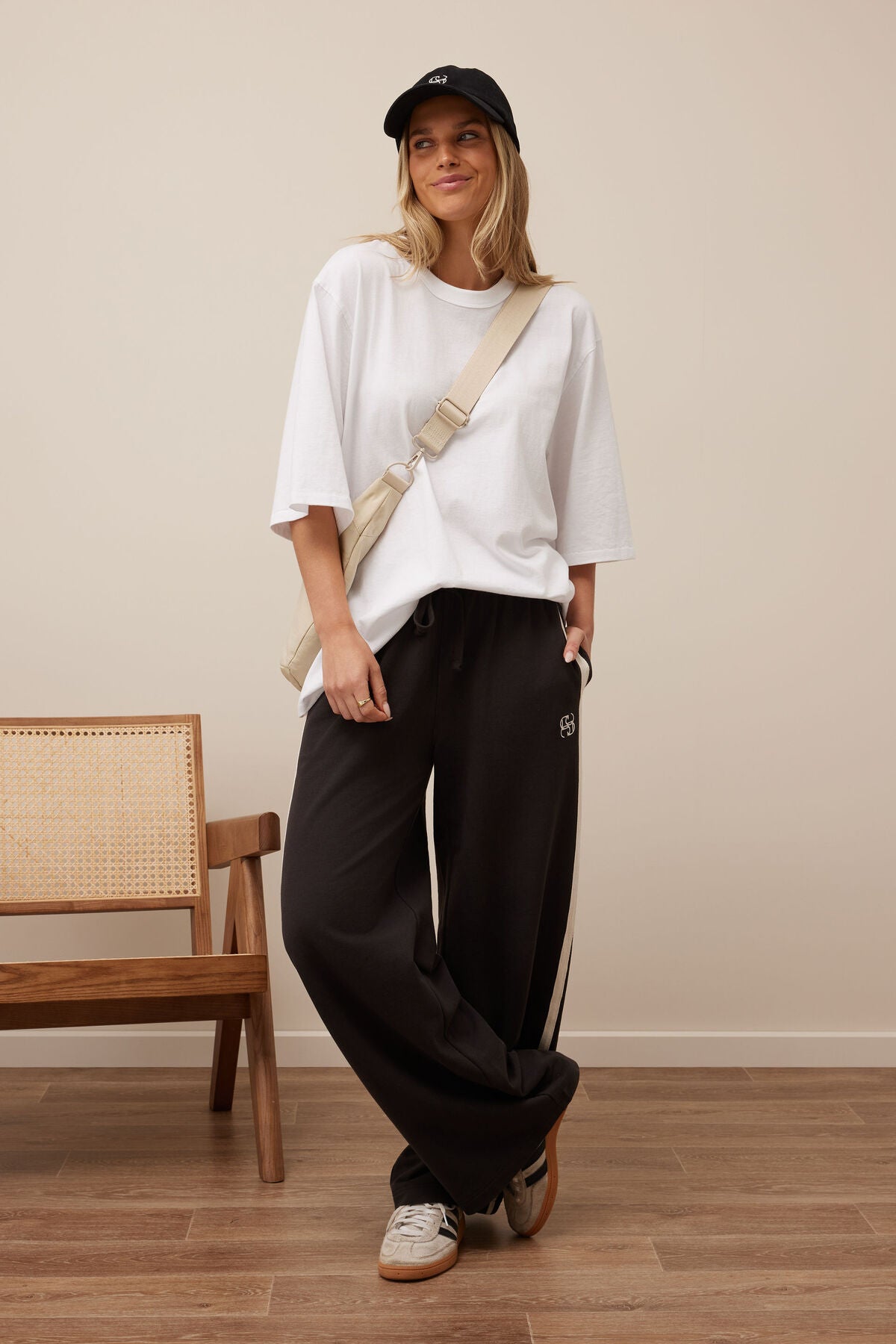 Woman wearing a white oversized shirt, Ceres Life black pants, and a cap, standing in a room with a wooden chair.