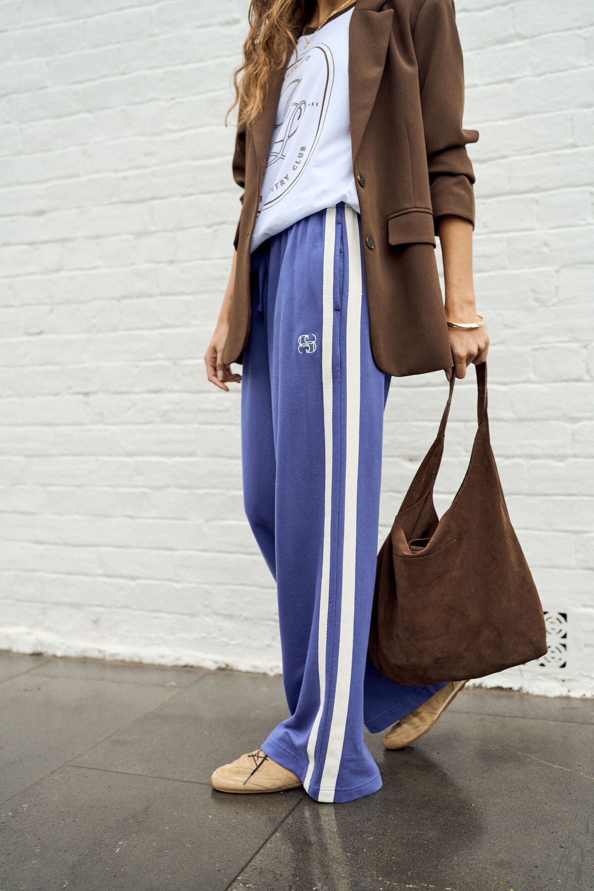 Image of A woman wearing wide-leg blue track pants with white side stripes, a brown blazer, a white graphic T-shirt, and tan loafers, carrying a brown suede hobo bag against a white brick wall.