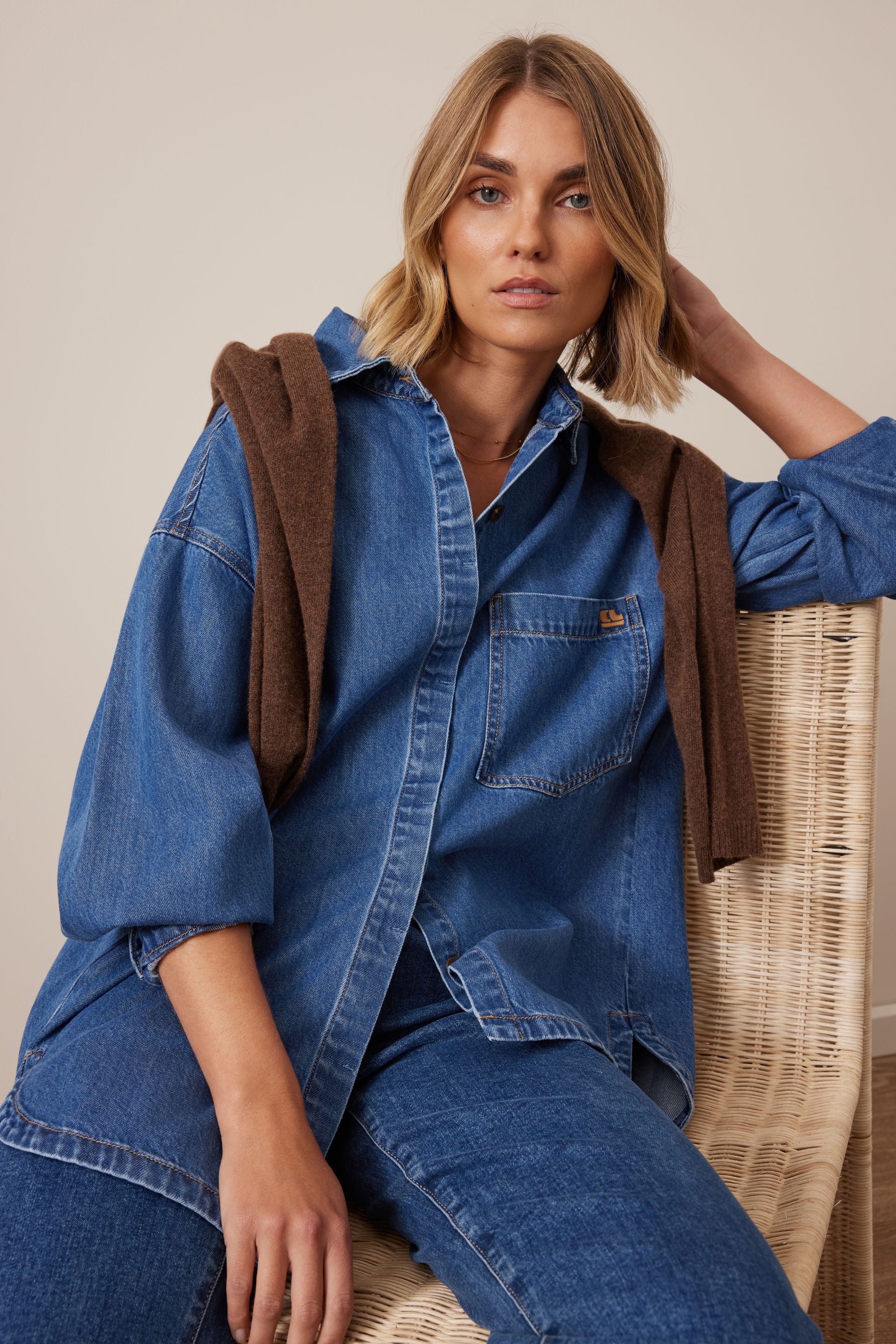 Woman wearing a Ceres Life blue denim outfit with a brown jacket, sitting on a wicker chair.
