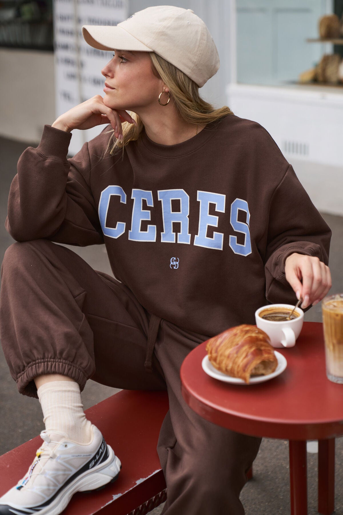 Person wearing a brown 'CERES' sweatshirt sitting at a small table with a croissant and coffee.