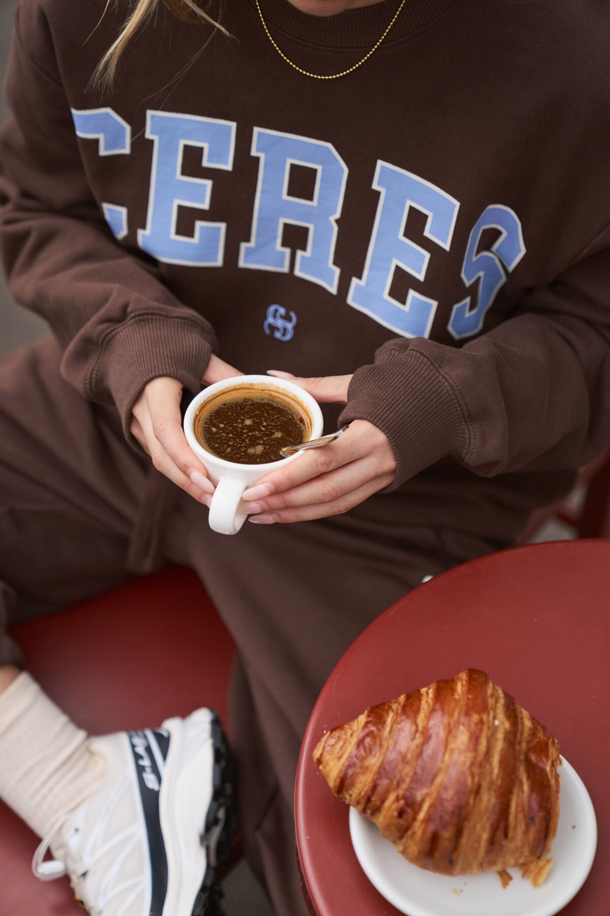 Woman wearing a brown sweatshirt with 'CERES' printed on it, holding a cup of coffee with a croissant on a plate.