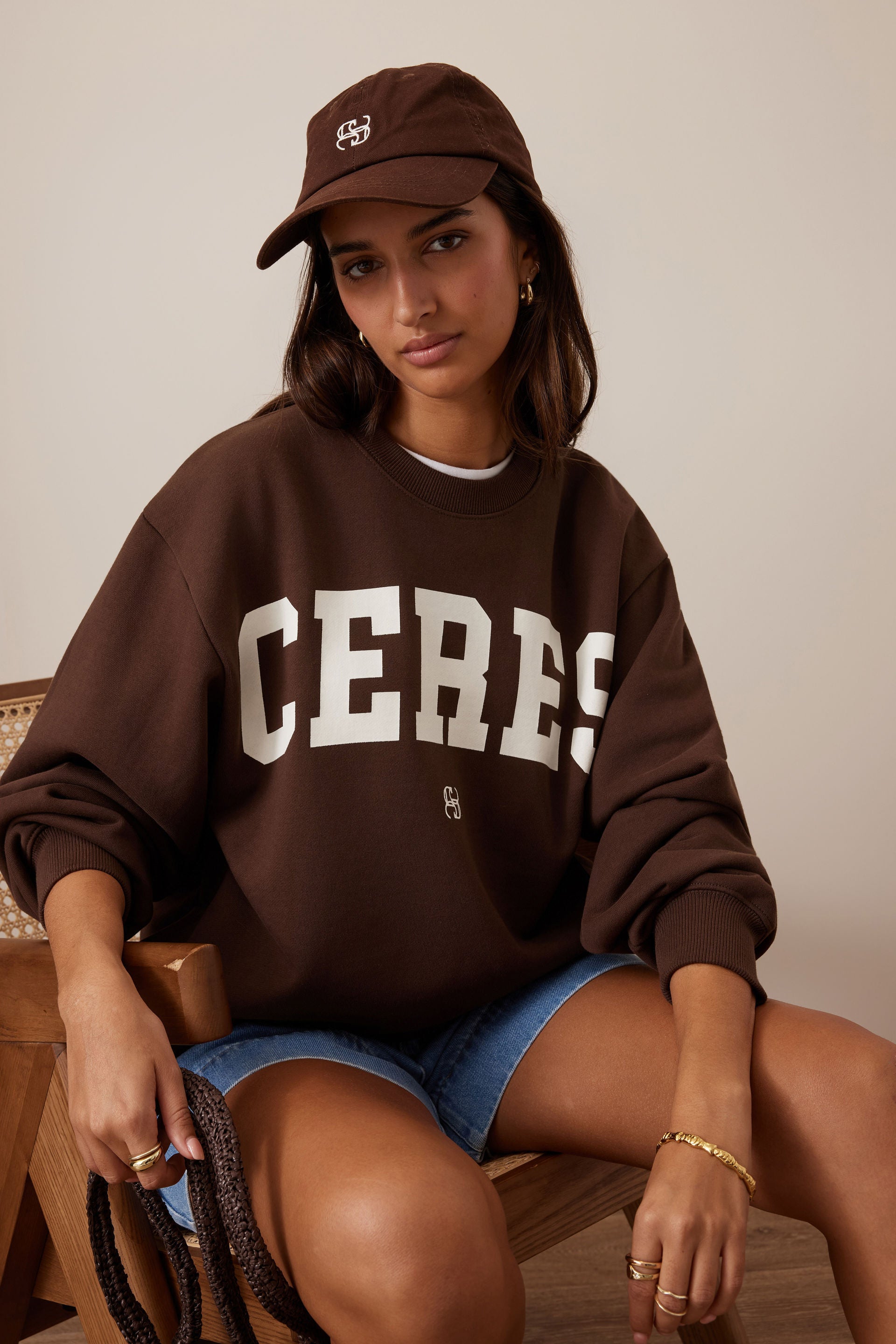 Image of Woman wearing a brown 'CERES' sweatshirt and denim shorts, sitting on a wooden chair.