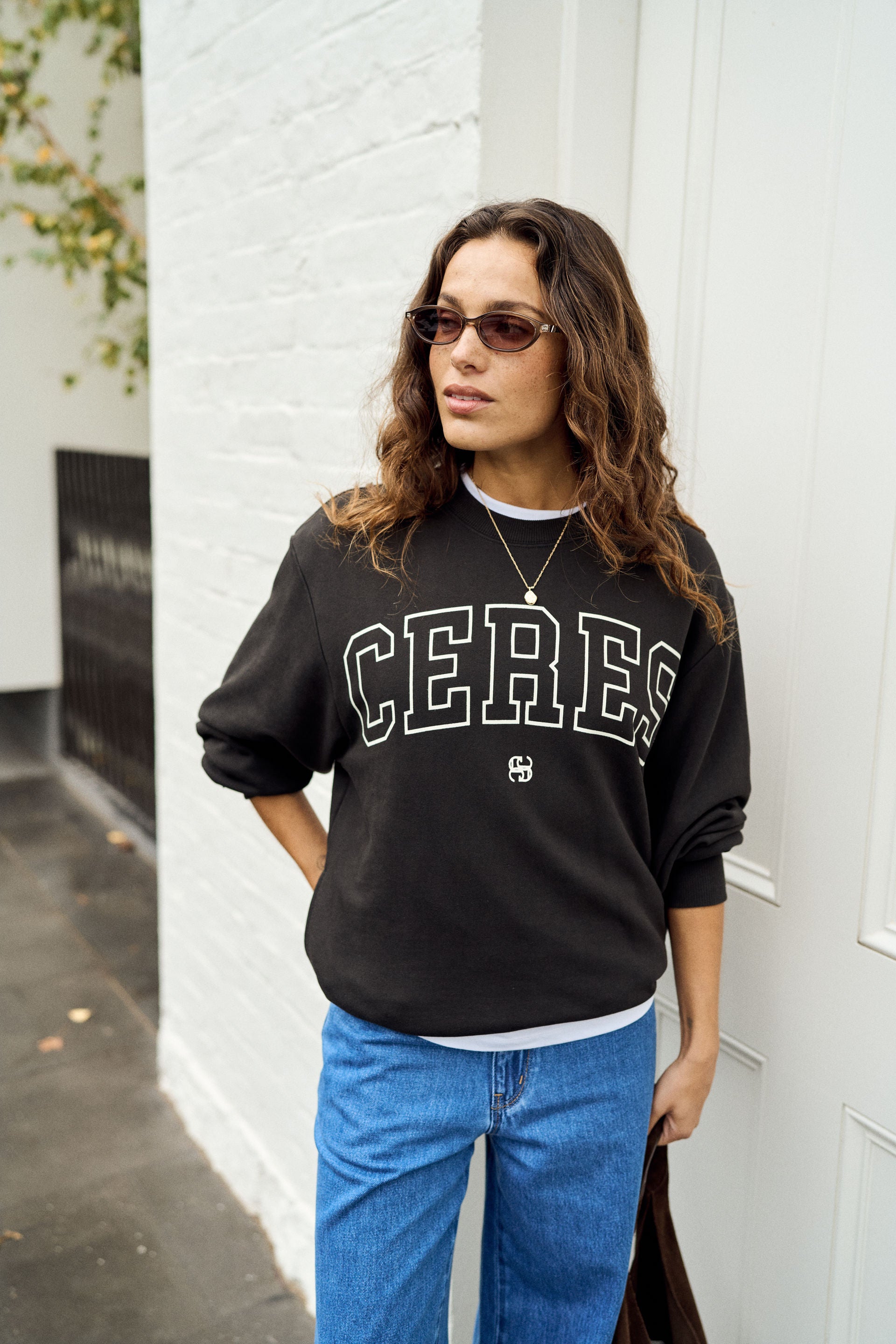 A woman wearing a black crewneck sweatshirt with 'CERES' printed in white collegiate-style letters across the chest, paired with blue jeans, standing against a white brick wall.