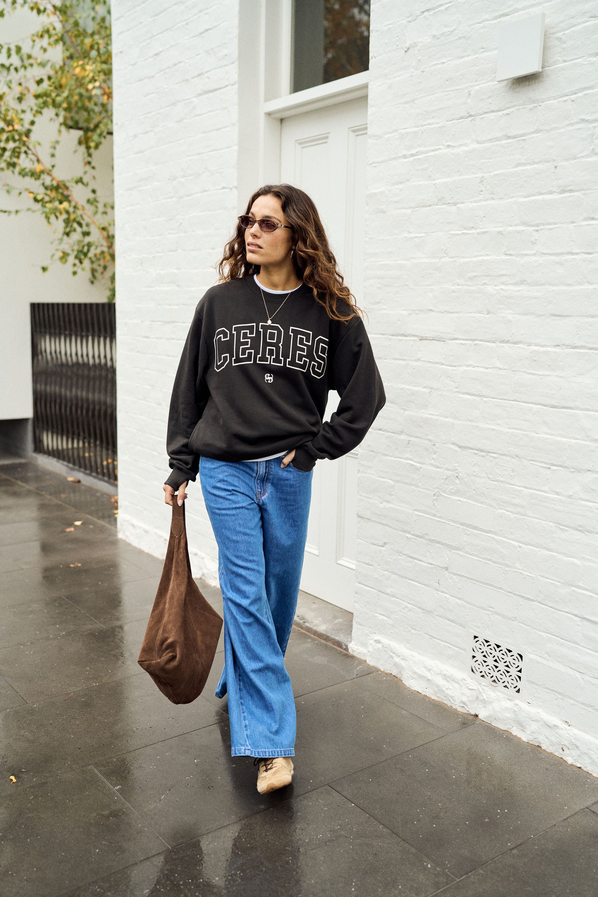 Image of A woman walking outdoors next to a white brick wall, wearing a black Ceres sweatshirt, blue wide-leg jeans, and carrying a brown leather tote bag.