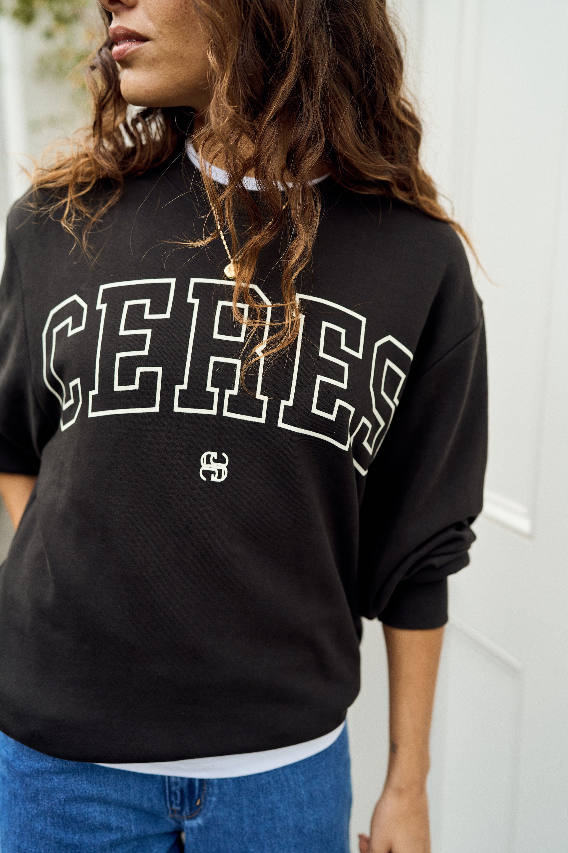 A woman wearing a black crewneck sweatshirt with 'CERES' printed in white varsity-style letters across the chest, paired with blue jeans.
