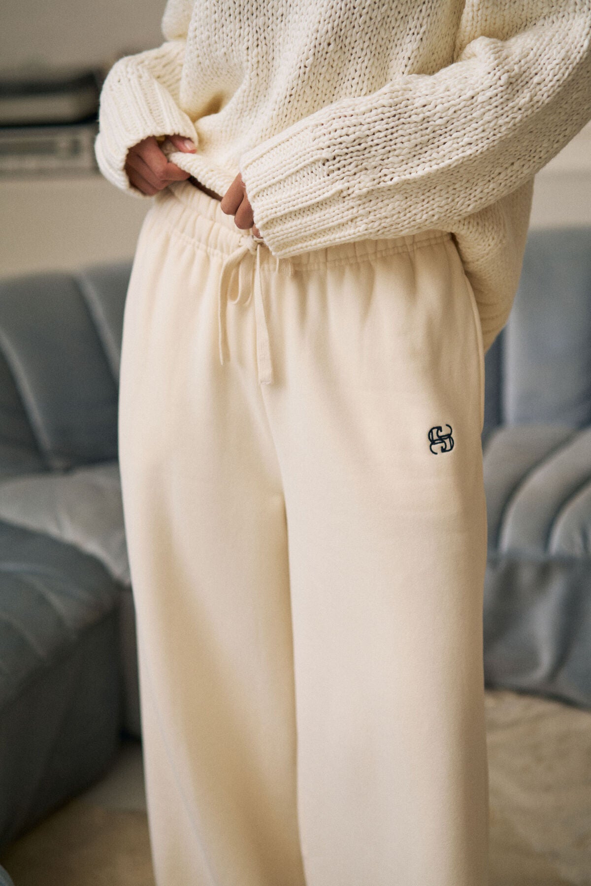 Image of Woman wearing a cream knitted sweater and Ceres Life beige pants with a logo, standing against a blurred indoor background.