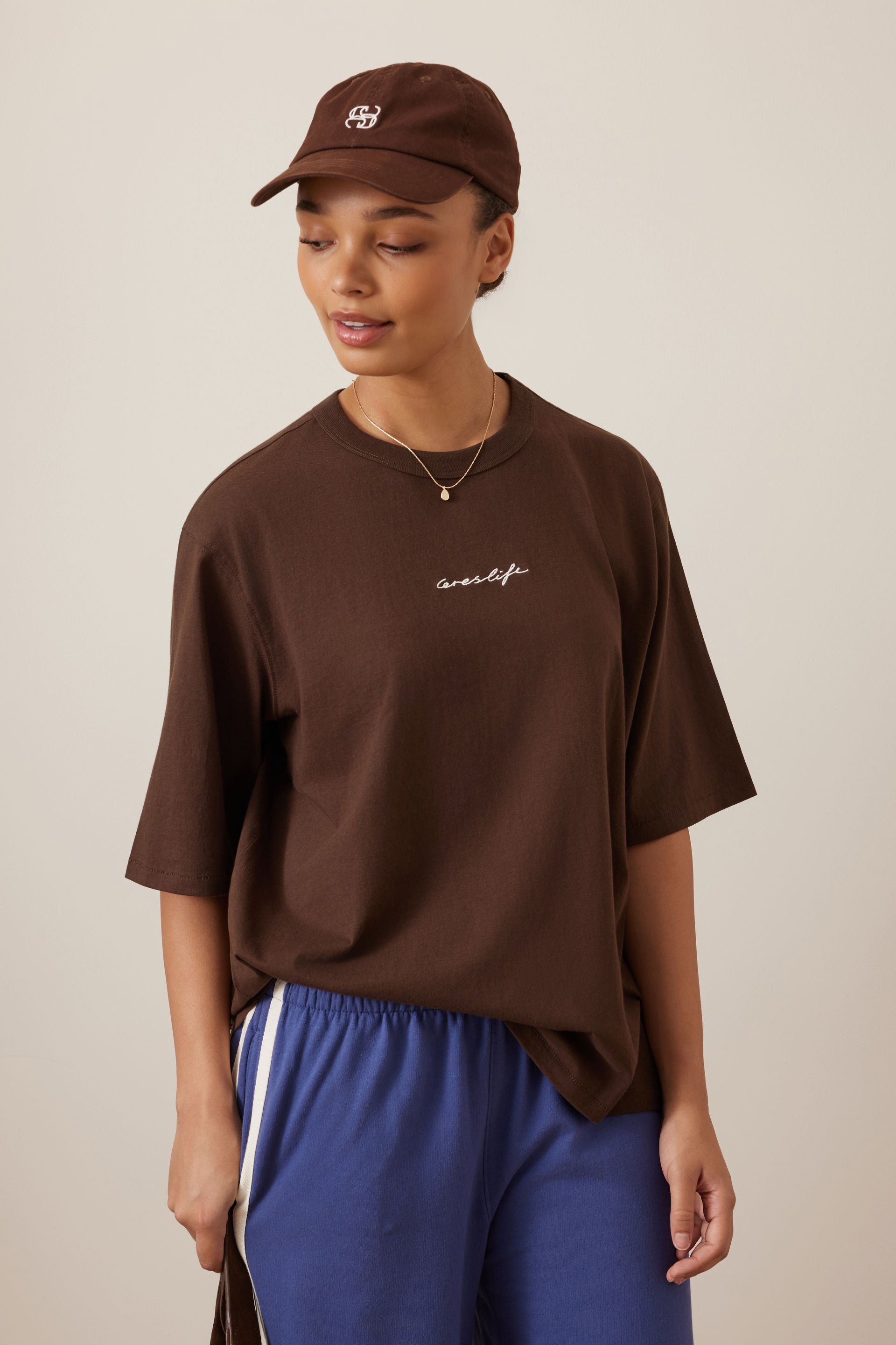 Image of A woman wearing a Ceres oversized brown t-shirt with a small white script logo on the chest, a matching brown baseball cap, and blue athletic pants, photographed against a neutral beige background.