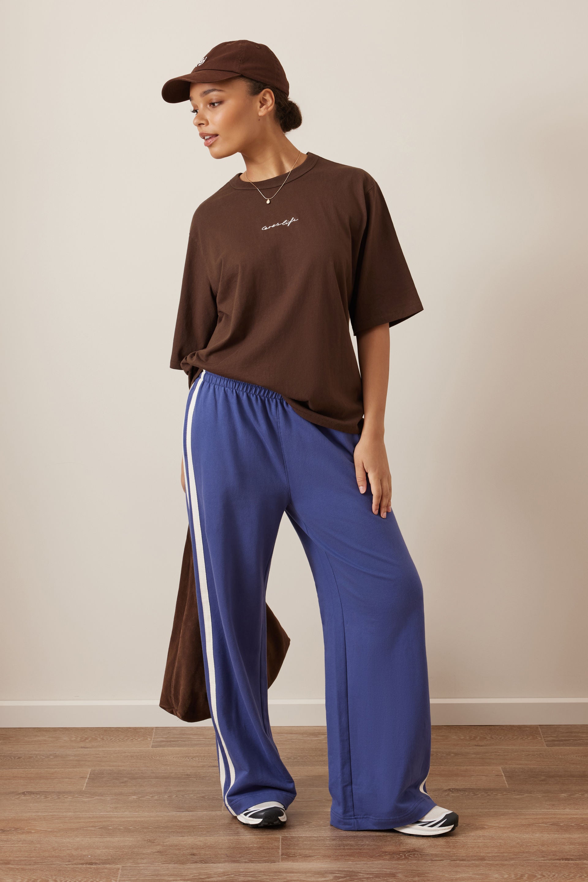 Image of A model wearing a Ceres brown t-shirt, blue wide-leg track pants with white side stripes, a brown cap, and sneakers, standing against a neutral beige wall and wooden floor.