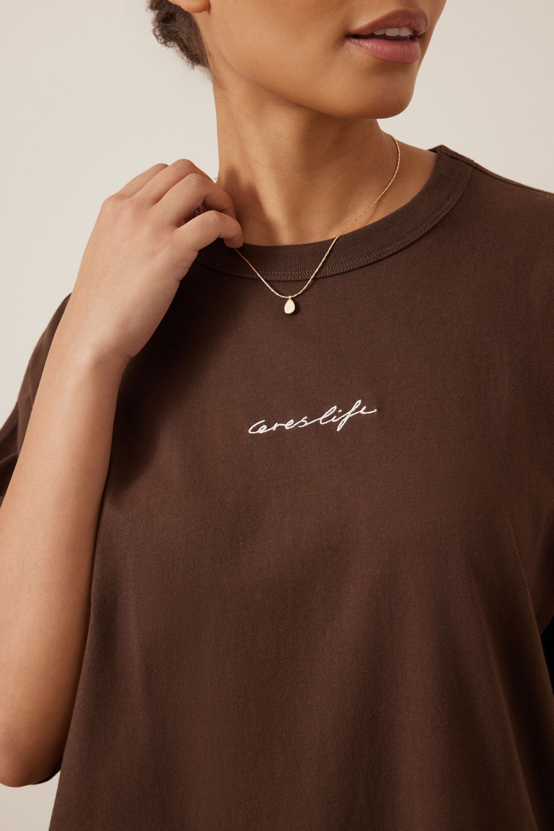 Image of A close-up of a woman wearing a Ceres Life brown crewneck t-shirt with 'cereslife' embroidered in white script on the chest, accessorized with a delicate gold necklace.