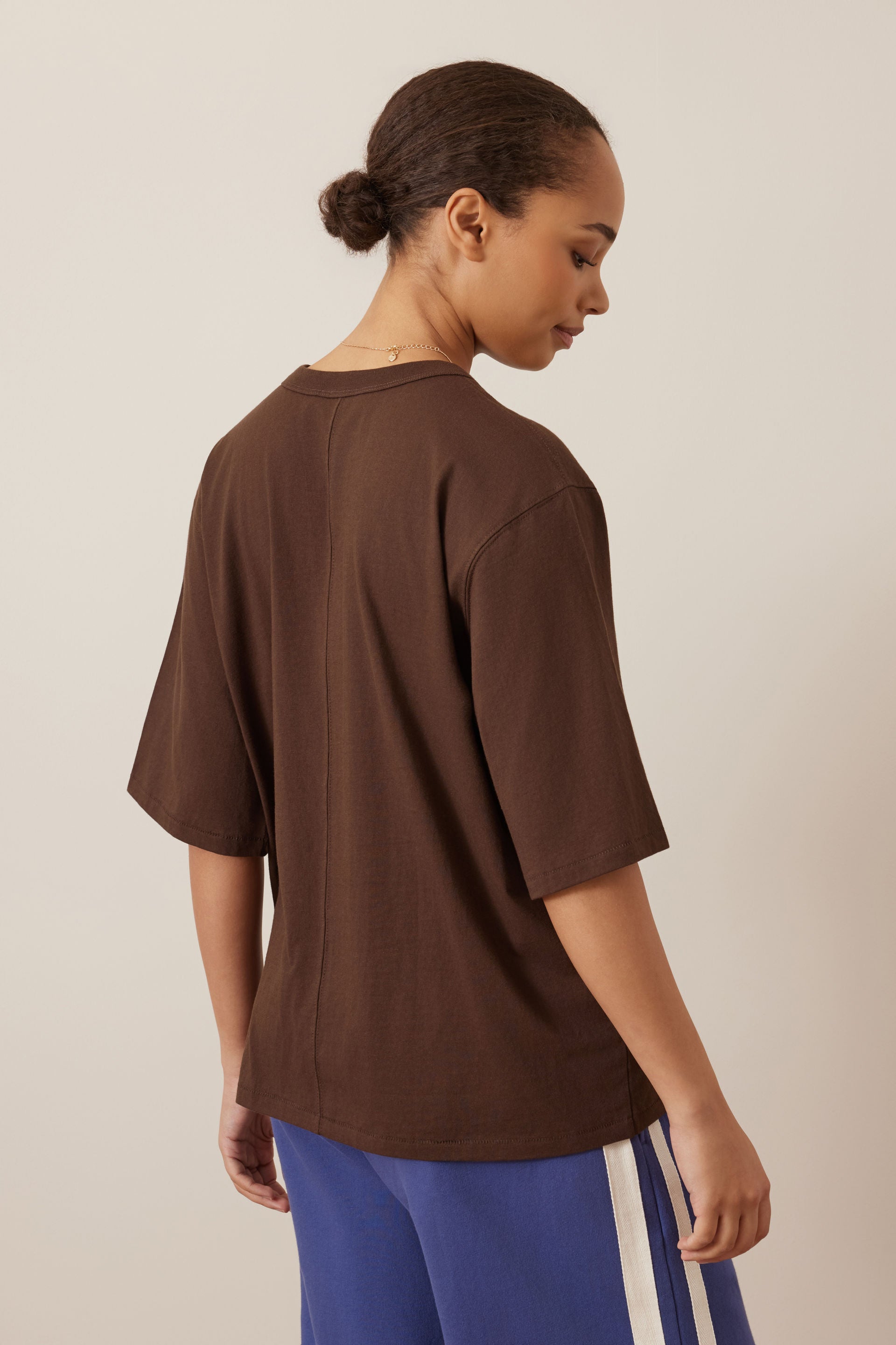A woman shown from the back wearing an oversized Ceres brown short-sleeve t-shirt and blue track pants with white side stripes, against a plain beige studio background.