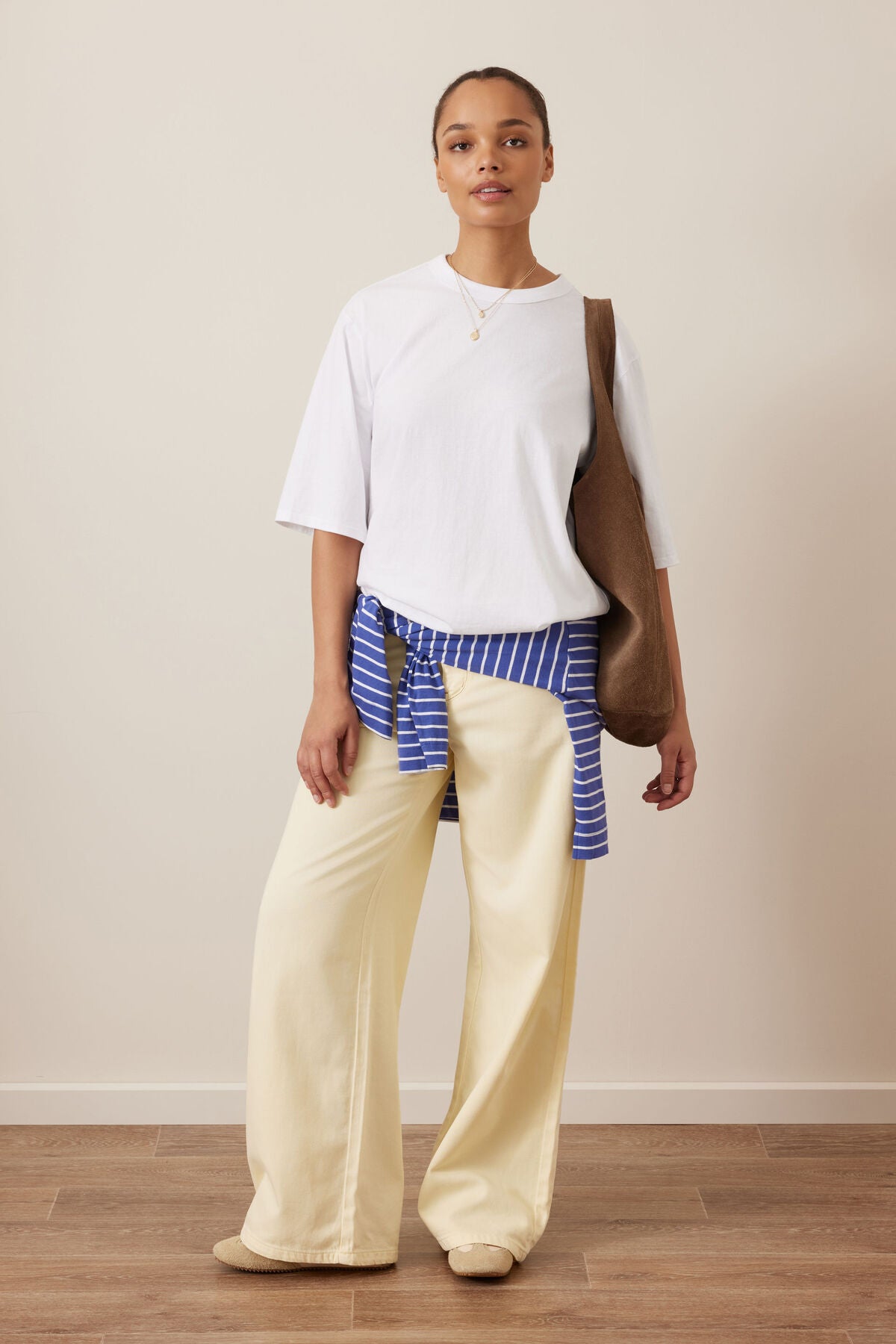 A model stands in a studio wearing a Ceres Life white short-sleeve top, cream wide-leg pants, a blue and white striped shirt tied around the waist, and a brown shoulder bag.
