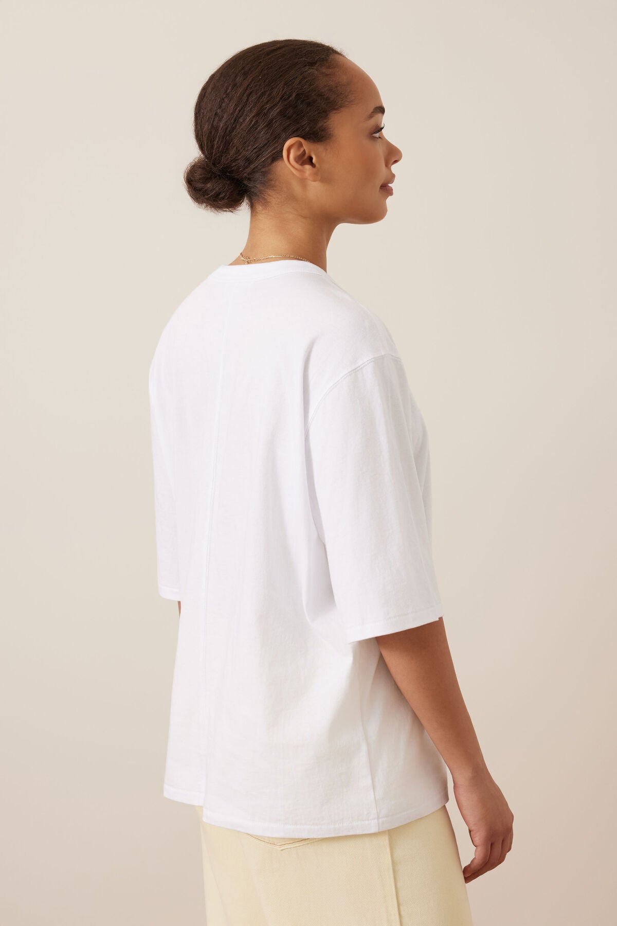 A woman shown from the back wearing an oversized Ceres Life white short-sleeve t-shirt and light beige pants, standing against a solid light beige background.
