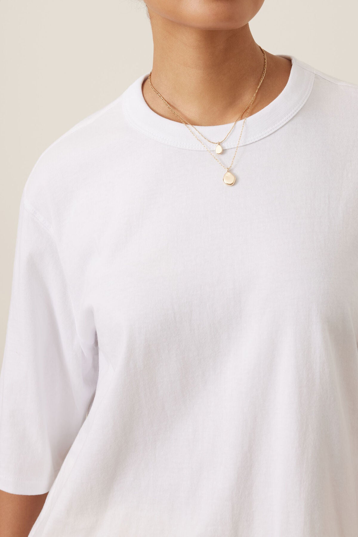 A close-up of a woman wearing a Ceres Life white short-sleeve crewneck t-shirt and layered gold necklaces with small disc pendants, set against a plain beige background.