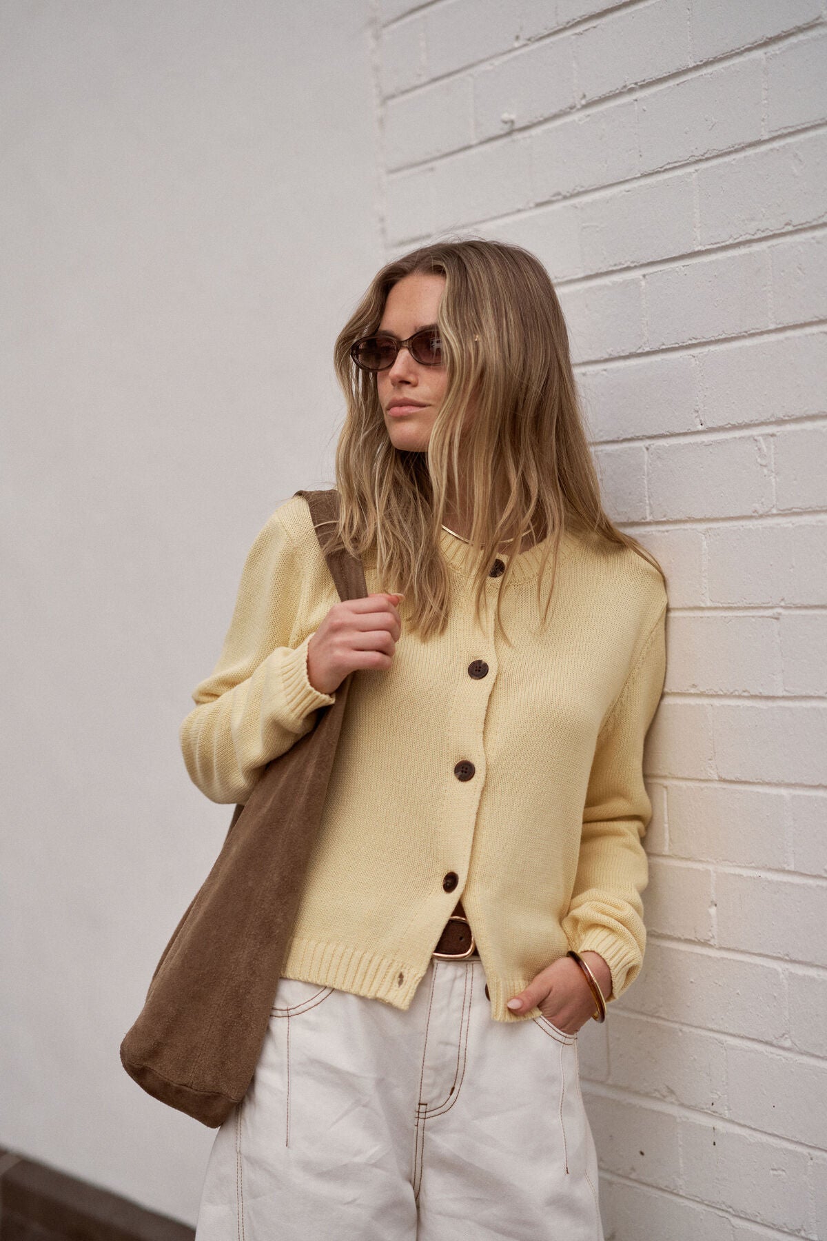 Image of Woman in a Ceres Life yellow cardigan holding a brown bag against a white brick wall.