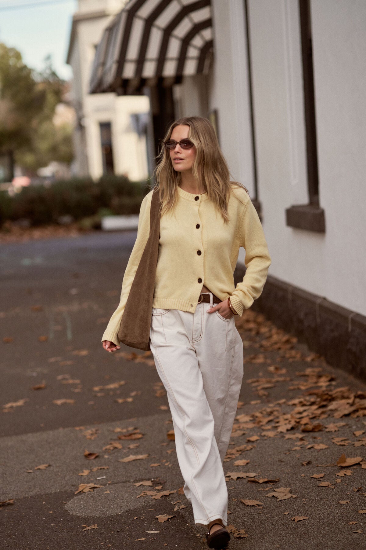 Woman in a Ceres Life yellow cardigan and white pants walking on a street with a building in the background.