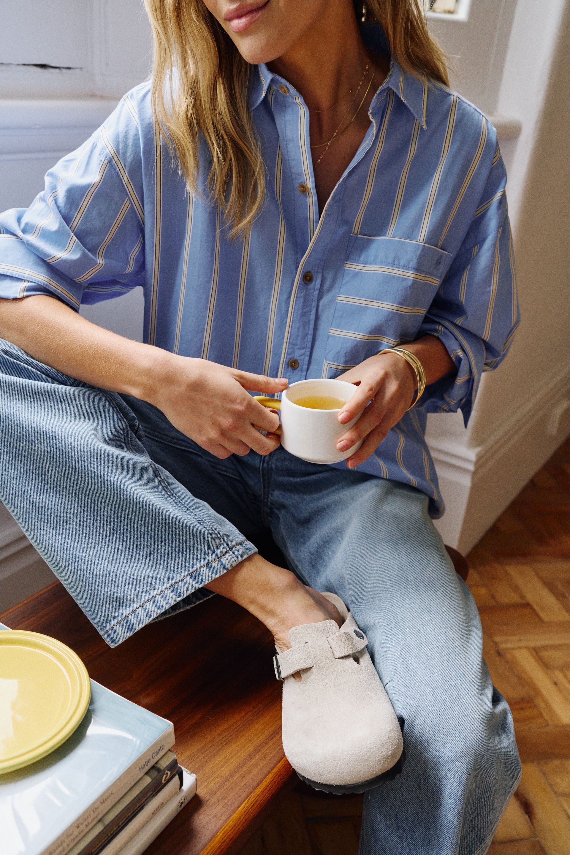 Image of A woman sitting indoors wearing a Ceres Life oversized light blue button-up shirt with thin yellow vertical stripes, light blue jeans, and off-white clog-style shoes, holding a white mug.