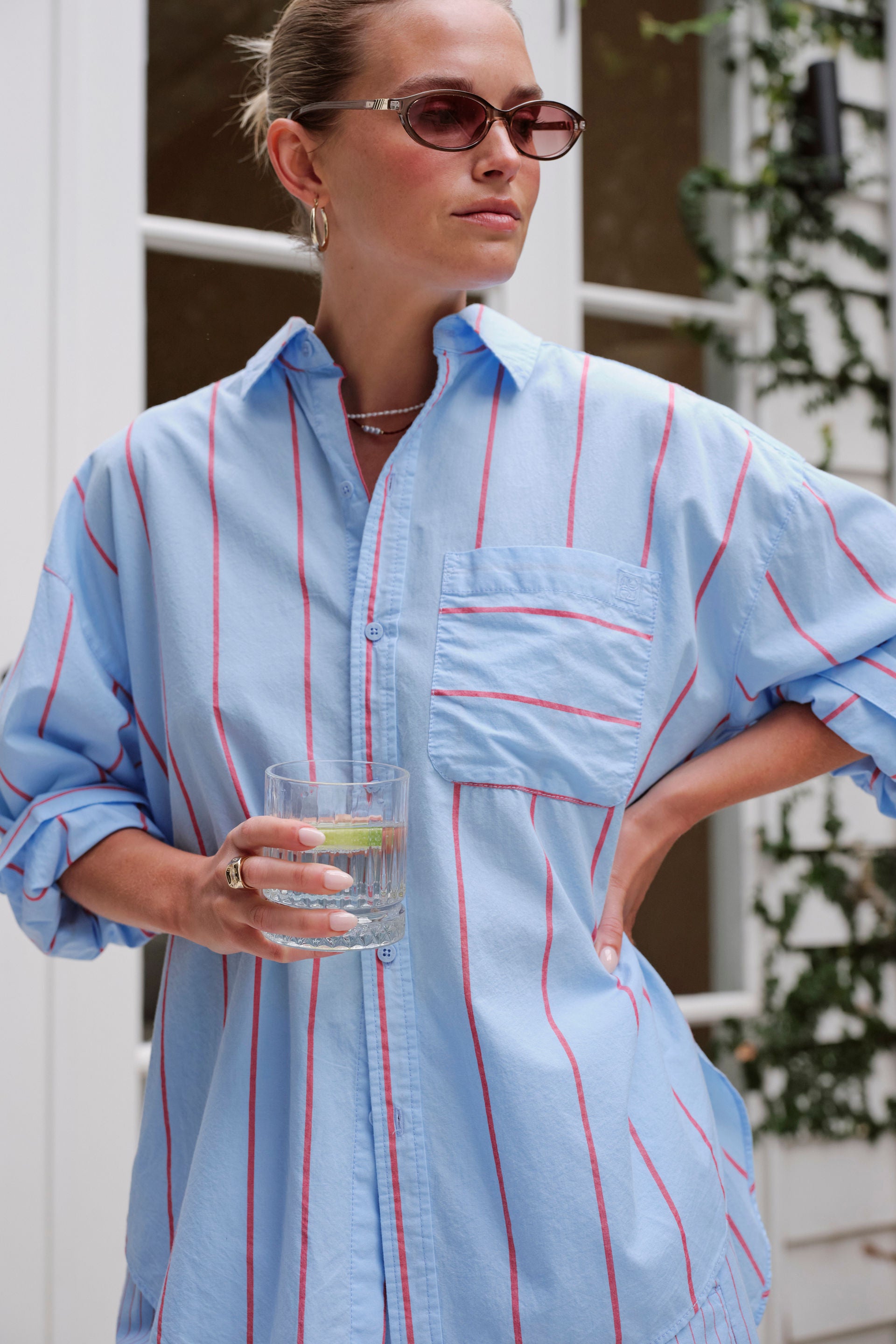 A woman wearing an oversized Ceres Light blue button-up shirt with thin red vertical stripes, holding a glass of water. She is wearing sunglasses and standing in front of a white window frame with green foliage.