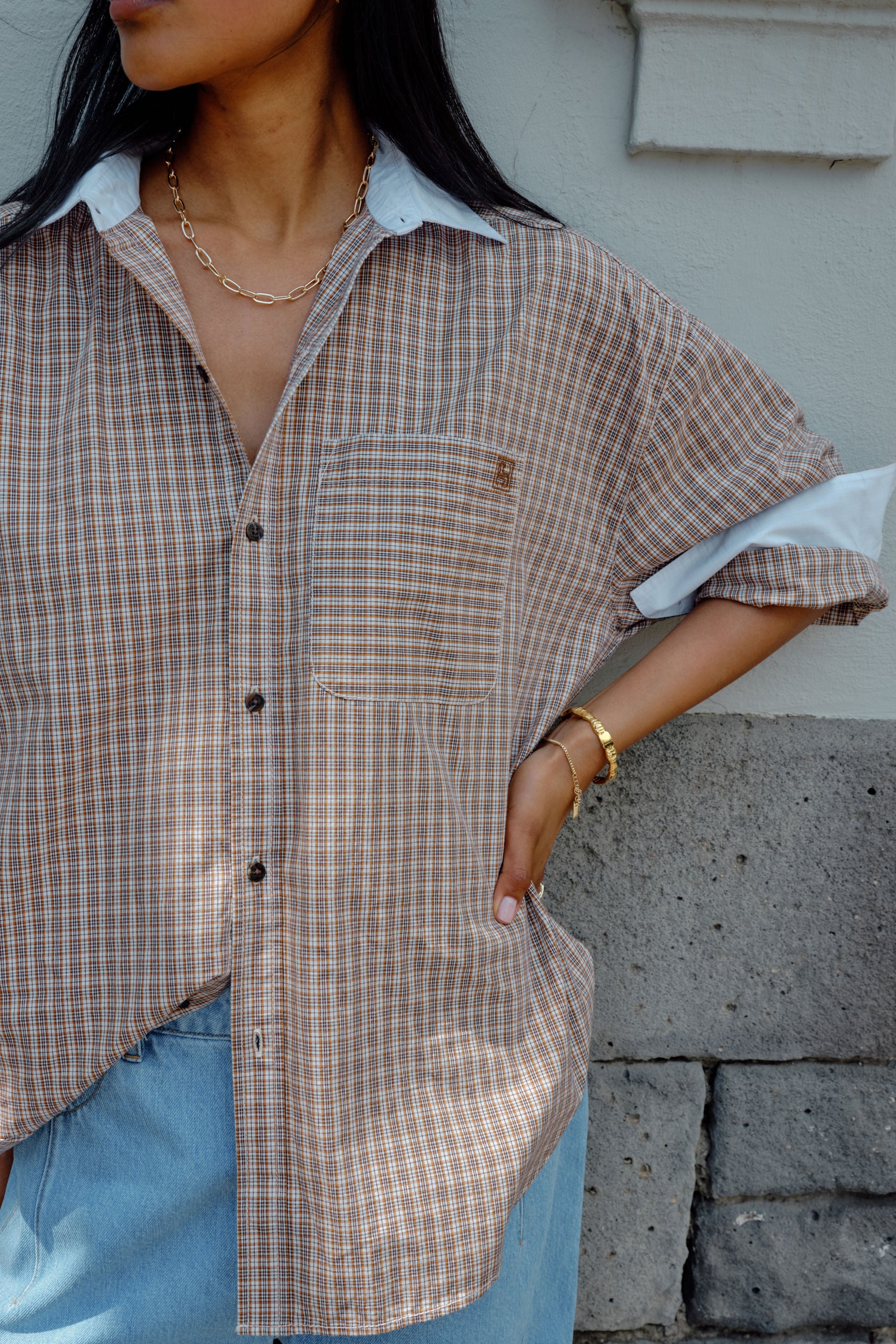 Woman wearing a Ceres Life plaid shirt and jeans with a brown bag, standing against a blue wall.