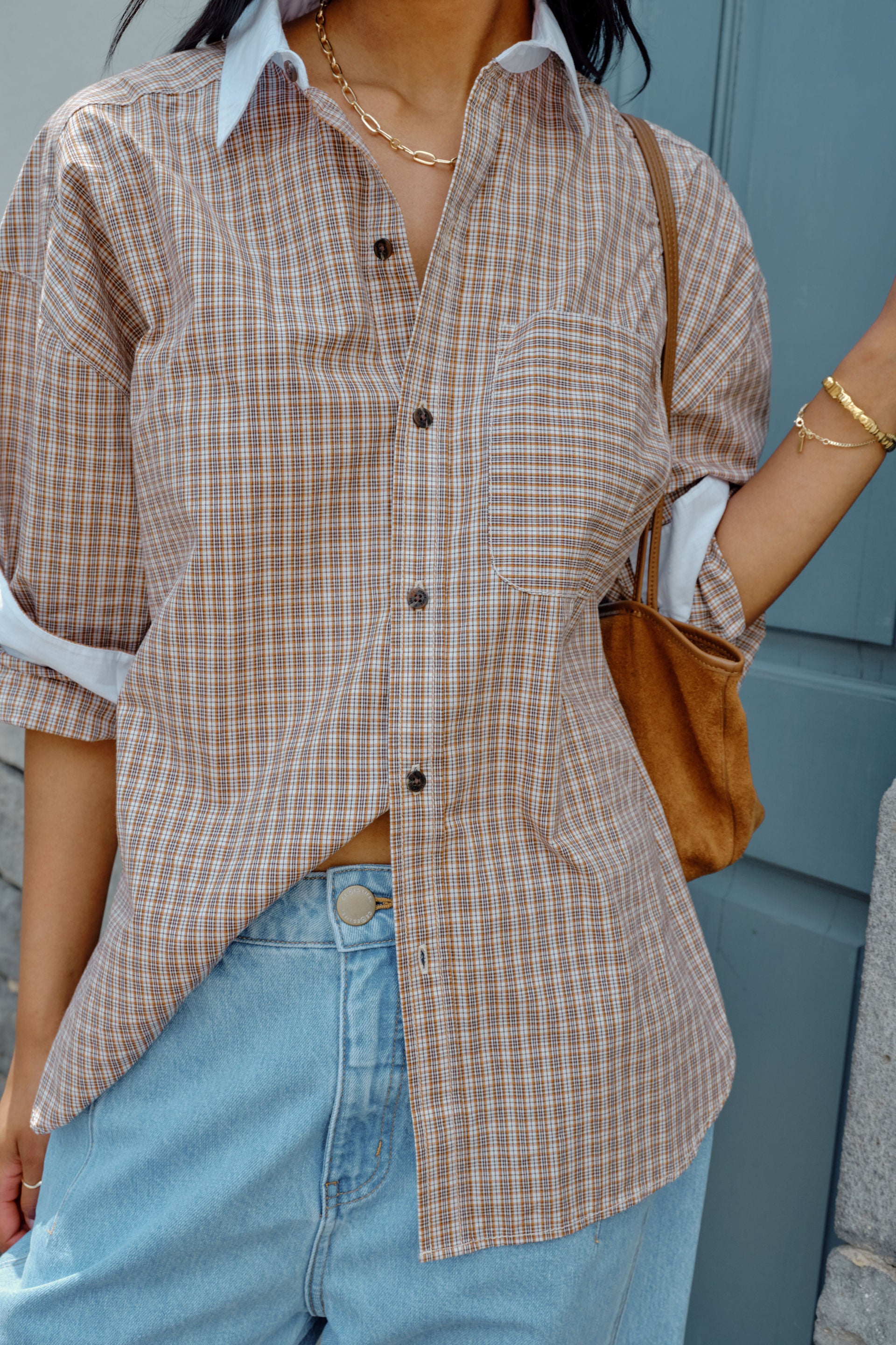 Image of Woman wearing a Ceres Life plaid shirt and jeans with a brown bag, standing against a blue wall.