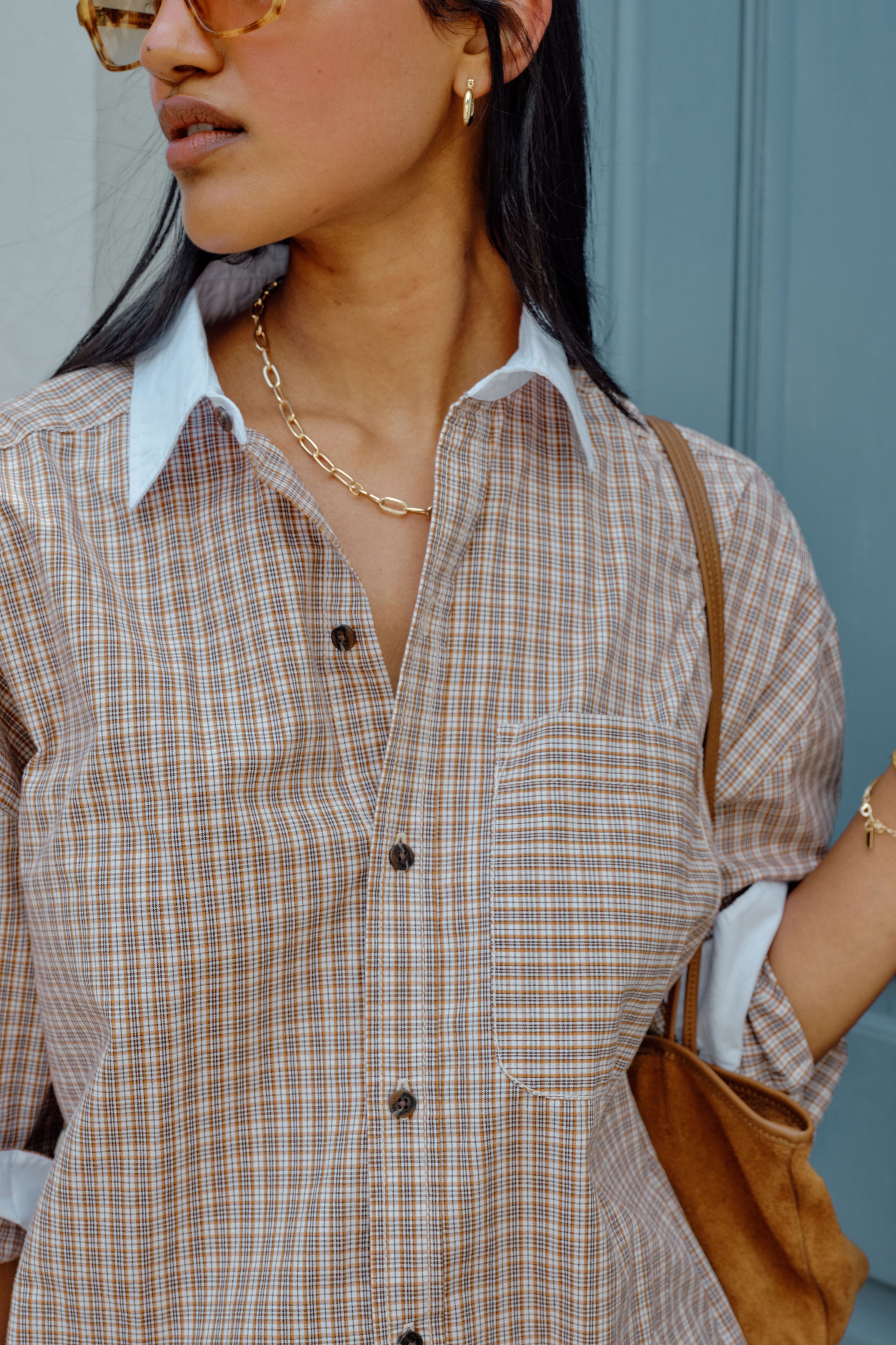 Image of Woman wearing a Ceres Life plaid shirt and jeans with a brown bag, standing against a blue wall.