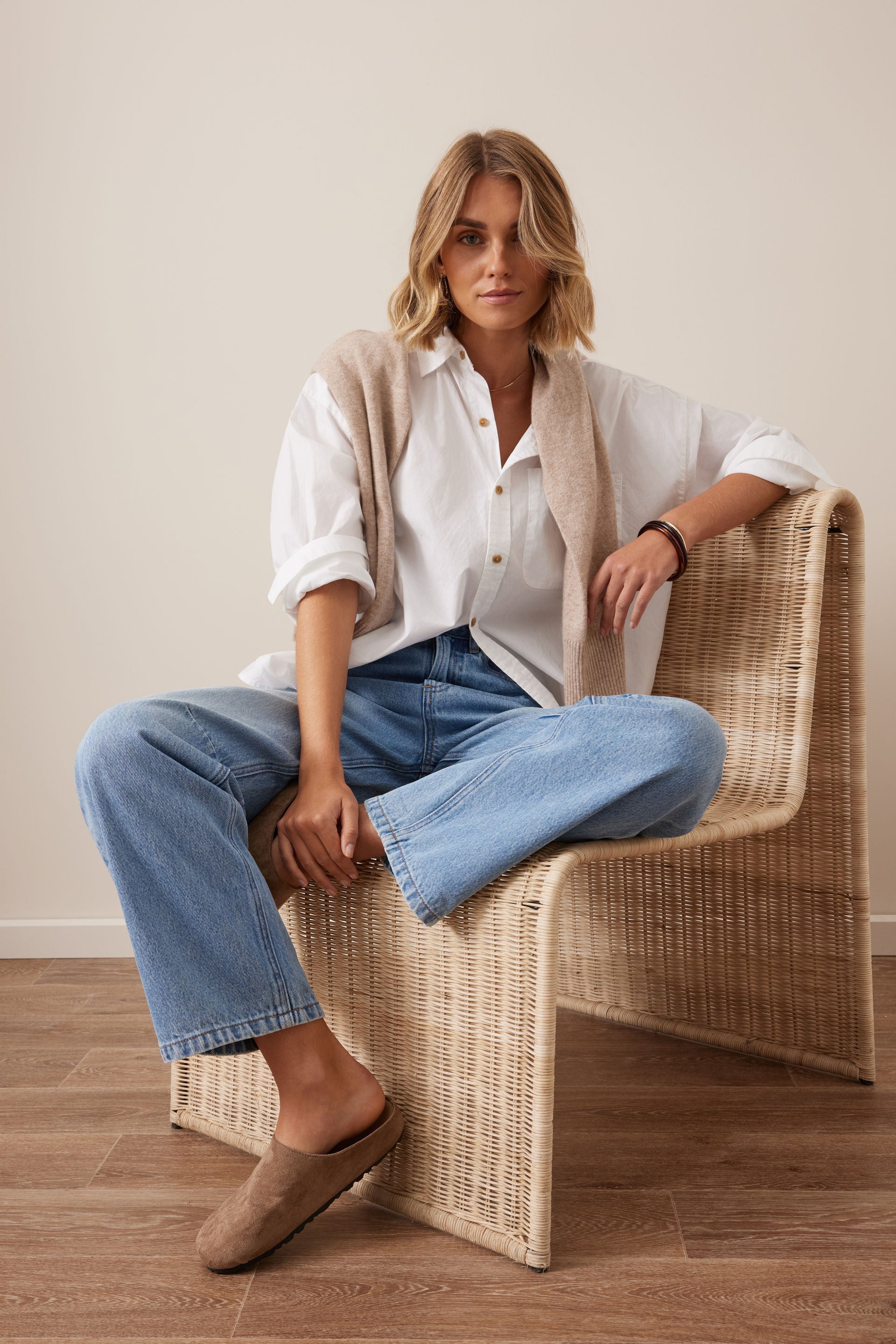 Image of Woman sitting on a wicker chair wearing a Ceres Life white shirt, beige cardigan, blue jeans, and brown loafers.