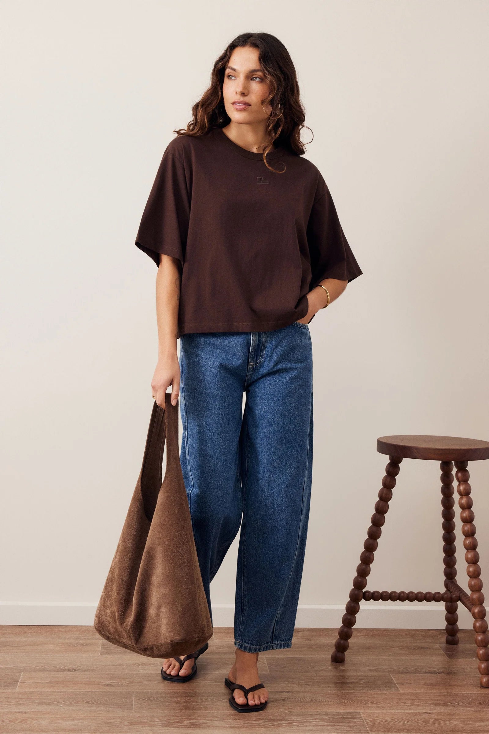 Image of Woman wearing a Ceres Life brown coloured tshirt with featured embroidery paired back with blue denim jeans on a plain background