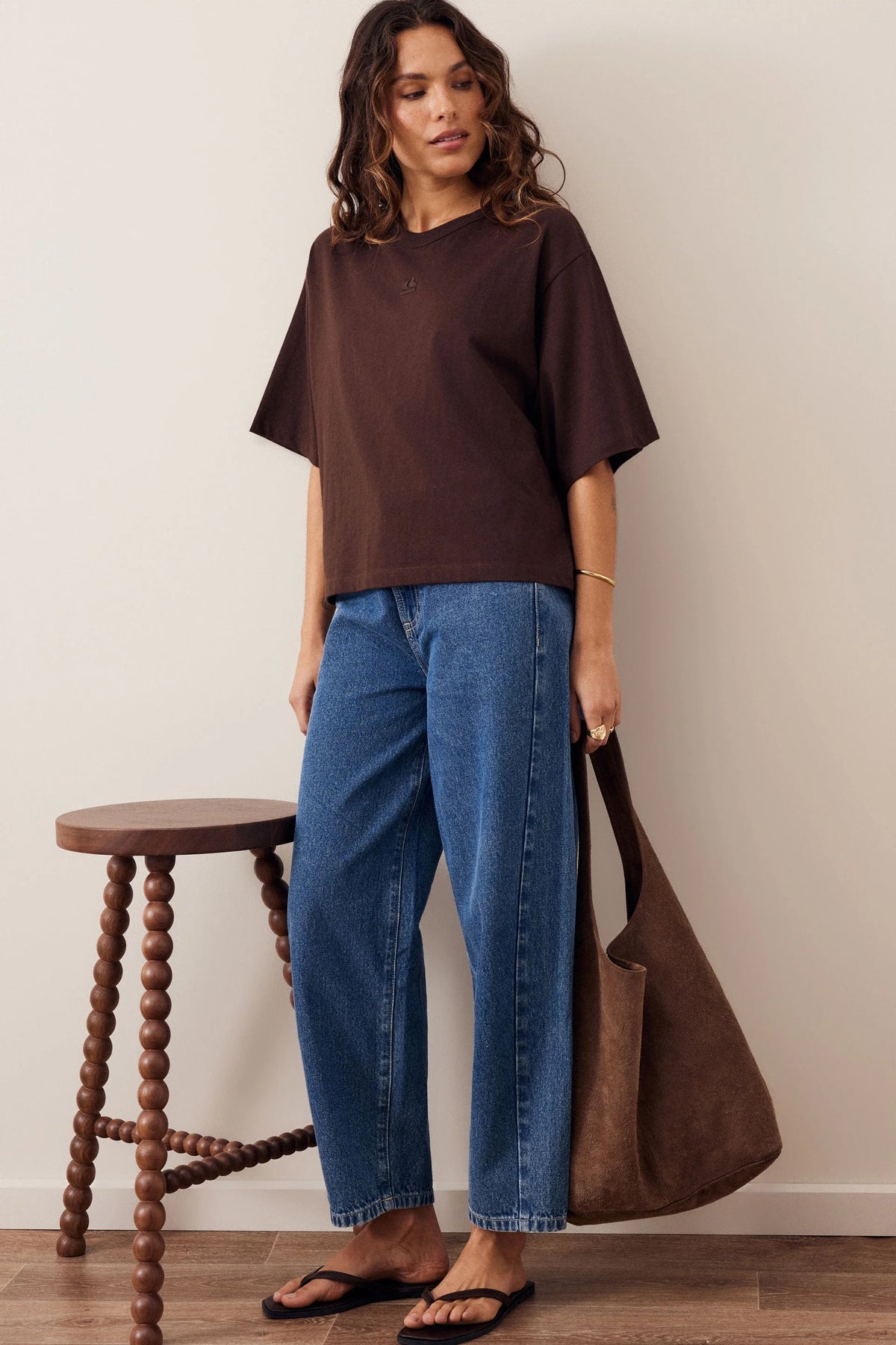 Woman wearing a Ceres Life brown coloured tshirt with featured embroidery paired back with blue denim jeans on a plain background