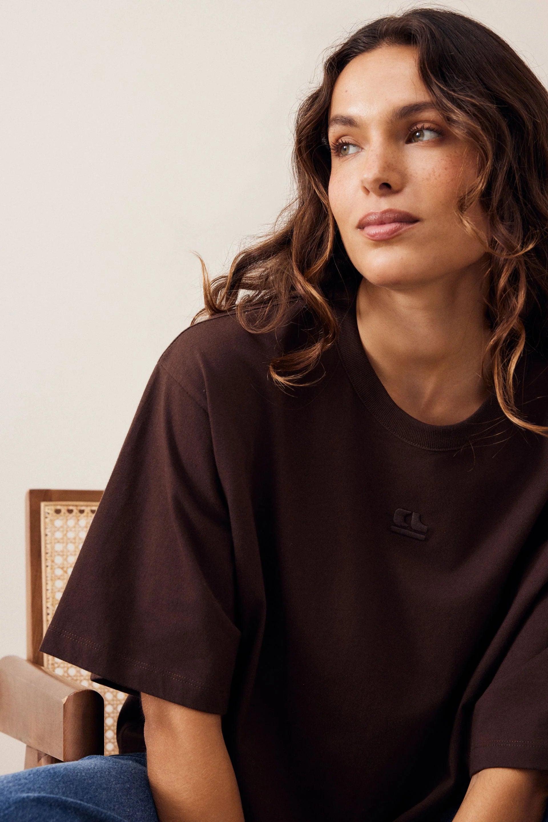 Image of Woman wearing a Ceres Life brown coloured tshirt with featured embroidery paired back with blue denim jeans on a plain background