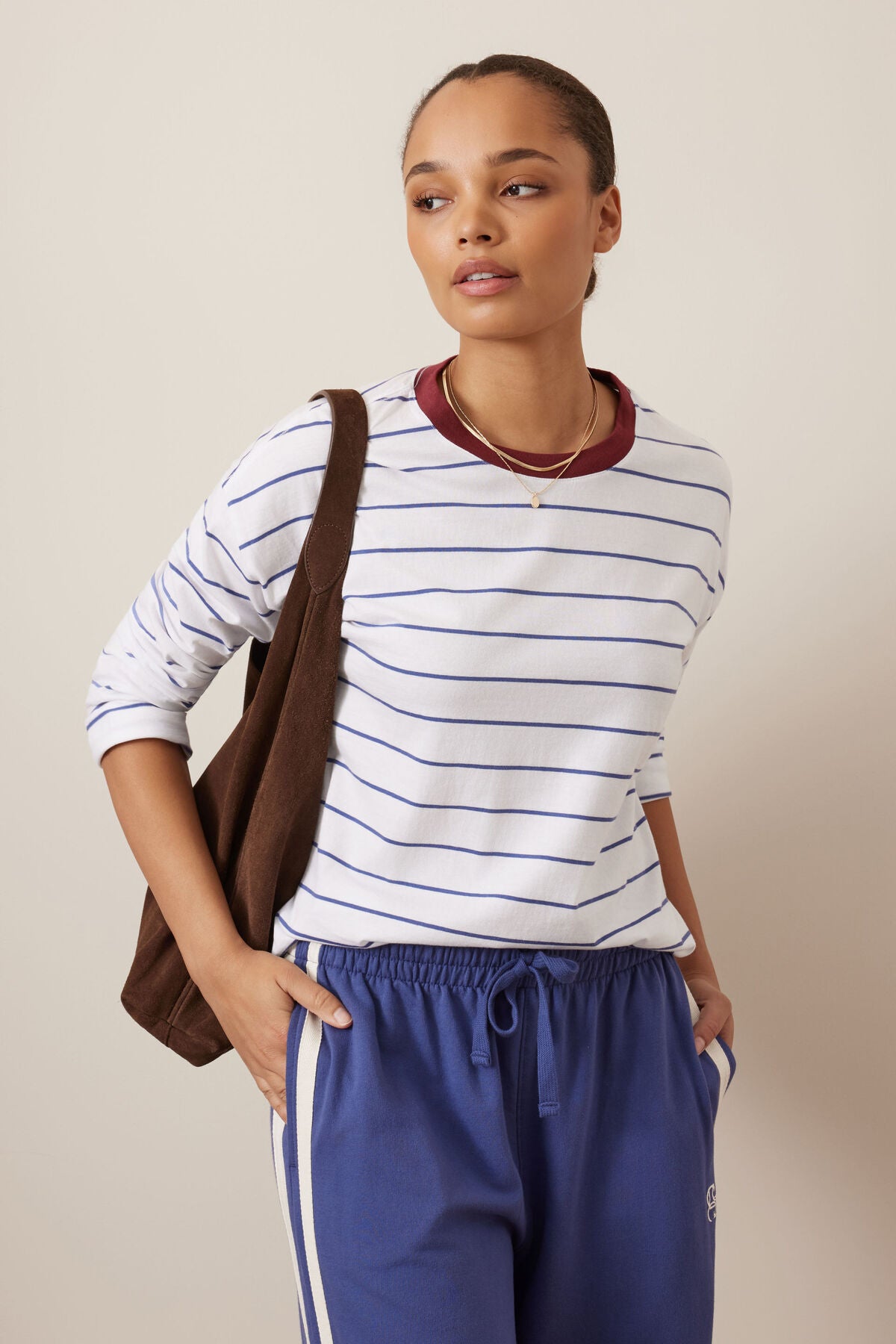 A woman modeling a Ceres Life white long-sleeve crewneck shirt with thin blue horizontal stripes and a dark red collar, paired with blue sweatpants and a brown shoulder bag, against a solid beige background.