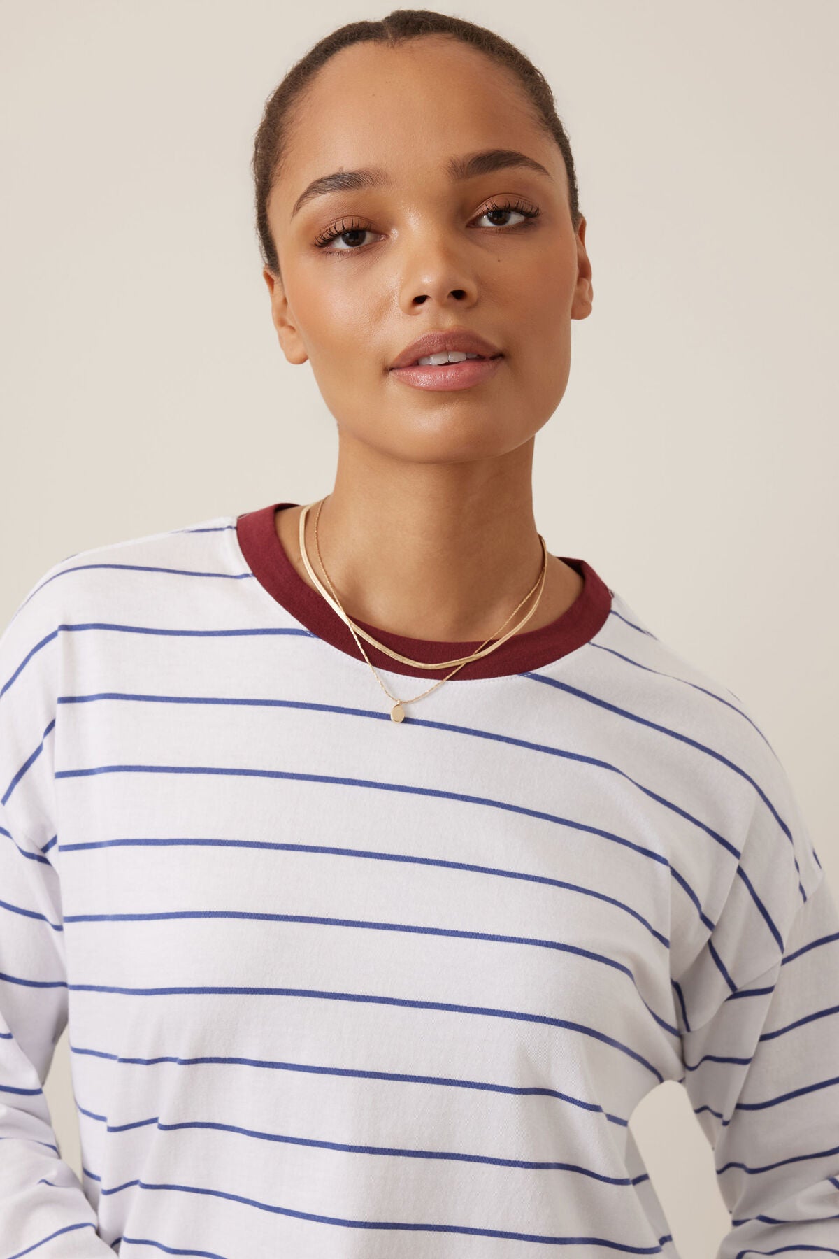 Image of Ceres Life white long-sleeve crewneck shirt with thin blue horizontal stripes and a contrasting dark red ribbed collar, styled with layered gold necklaces against a plain beige background.