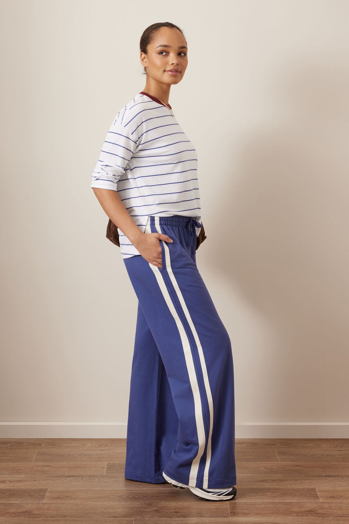 A woman modeling a Ceres Life white long-sleeve top with thin navy horizontal stripes and blue wide-leg pants featuring two white side stripes, standing in a studio with a beige wall and wood floor.