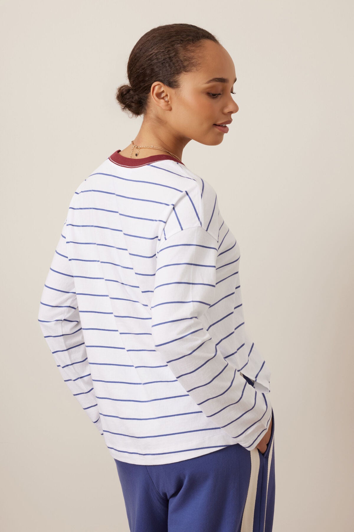 A woman shown from the back and side wearing a Ceres Life white long-sleeve top with thin blue horizontal stripes and a dark red crew neck, paired with blue pants, against a plain beige studio background.