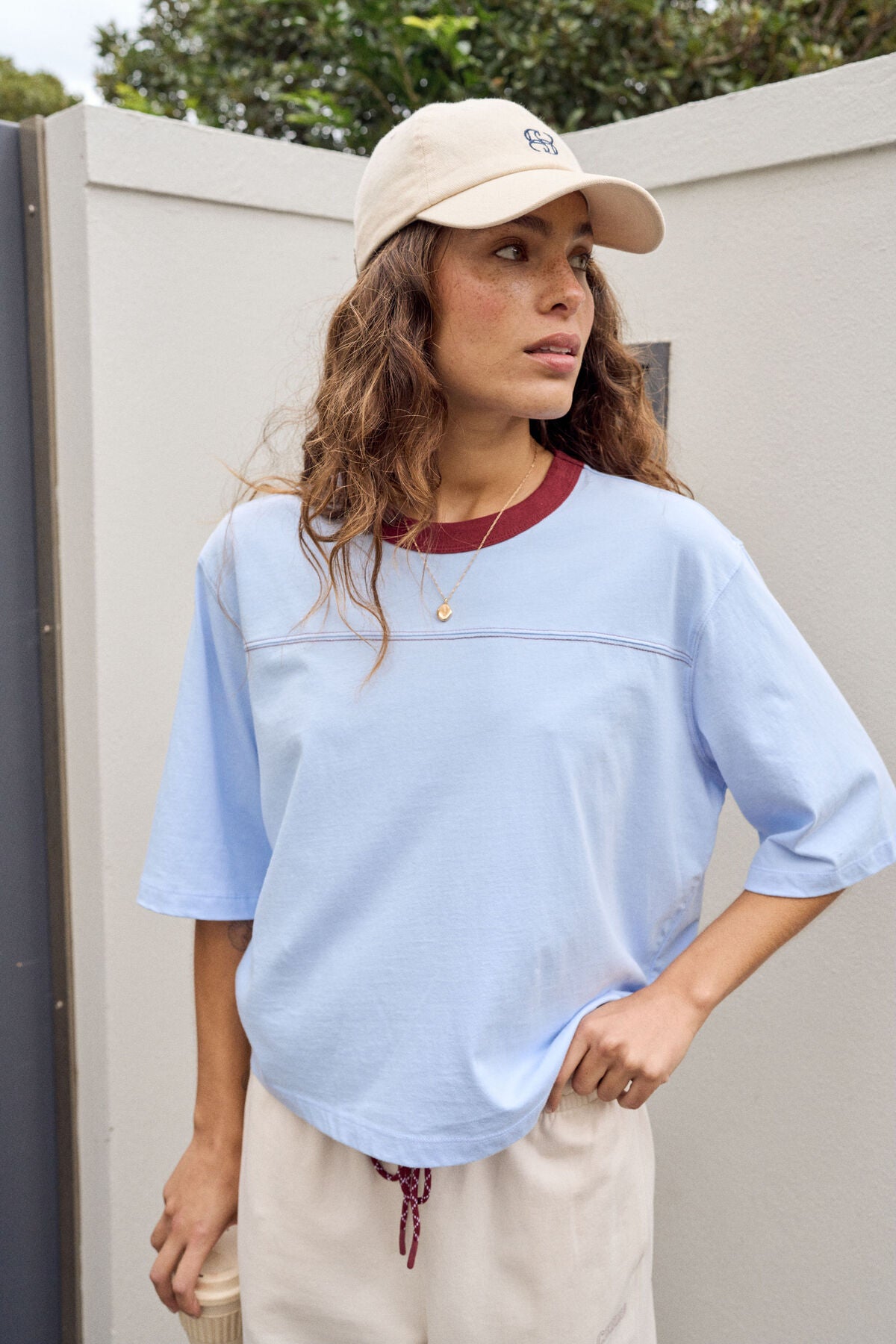 A woman wearing a Ceres Life light blue short-sleeve crop top, cream wide-leg pants, and a beige baseball cap stands outdoors next to a white wall and a dark gray gate, holding a takeaway coffee cup.