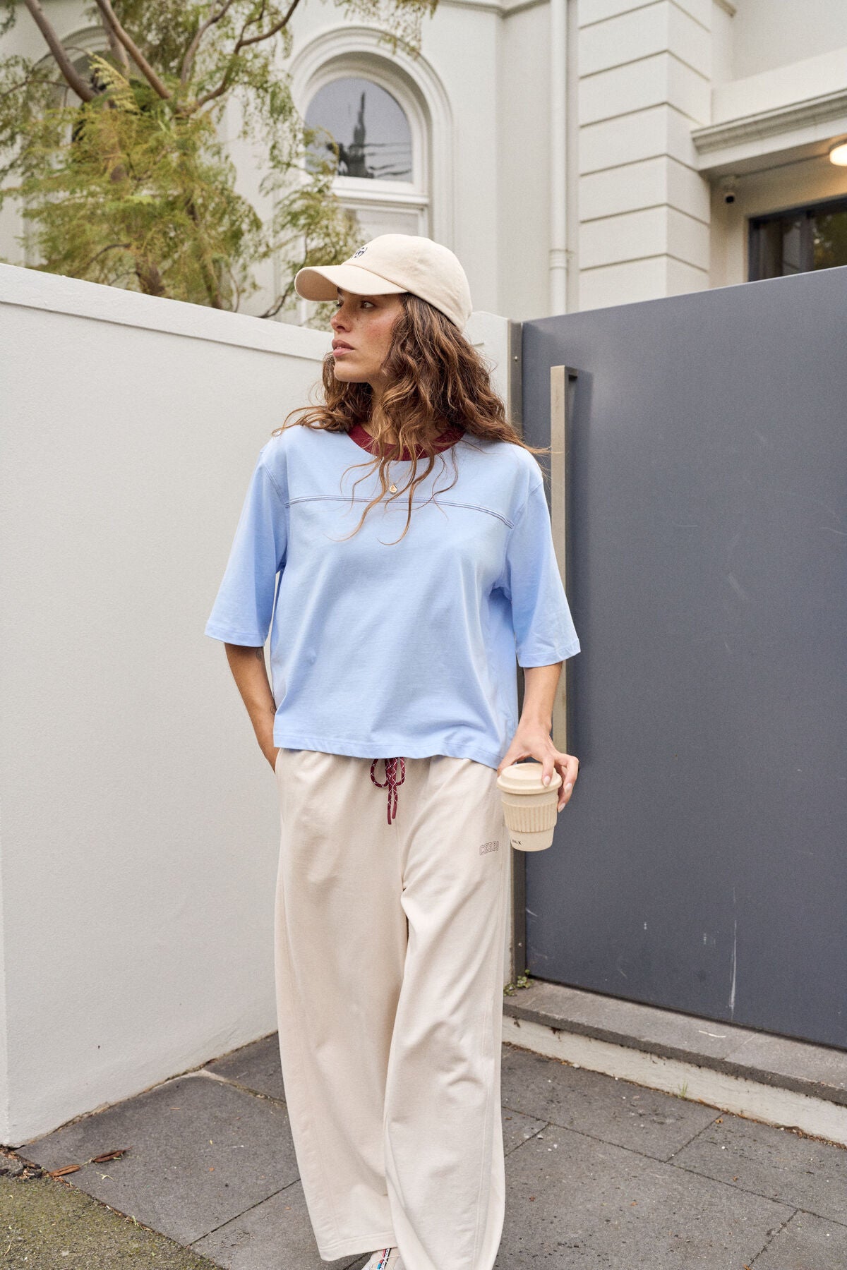 Image of A woman wearing a Ceres Life light blue short-sleeve crop top, cream wide-leg pants, and a beige baseball cap stands outdoors next to a white wall and a dark gray gate, holding a takeaway coffee cup.