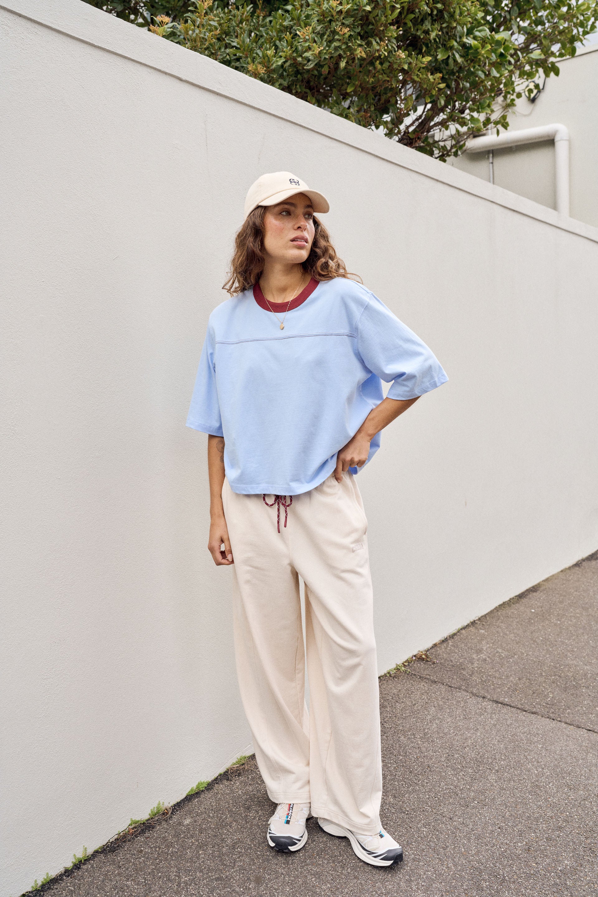 Image of A woman stands outdoors against a white wall wearing a Ceres Life light blue oversized short-sleeve top with a dark red collar, cream drawstring wide-leg pants, white sneakers, and a beige baseball cap.