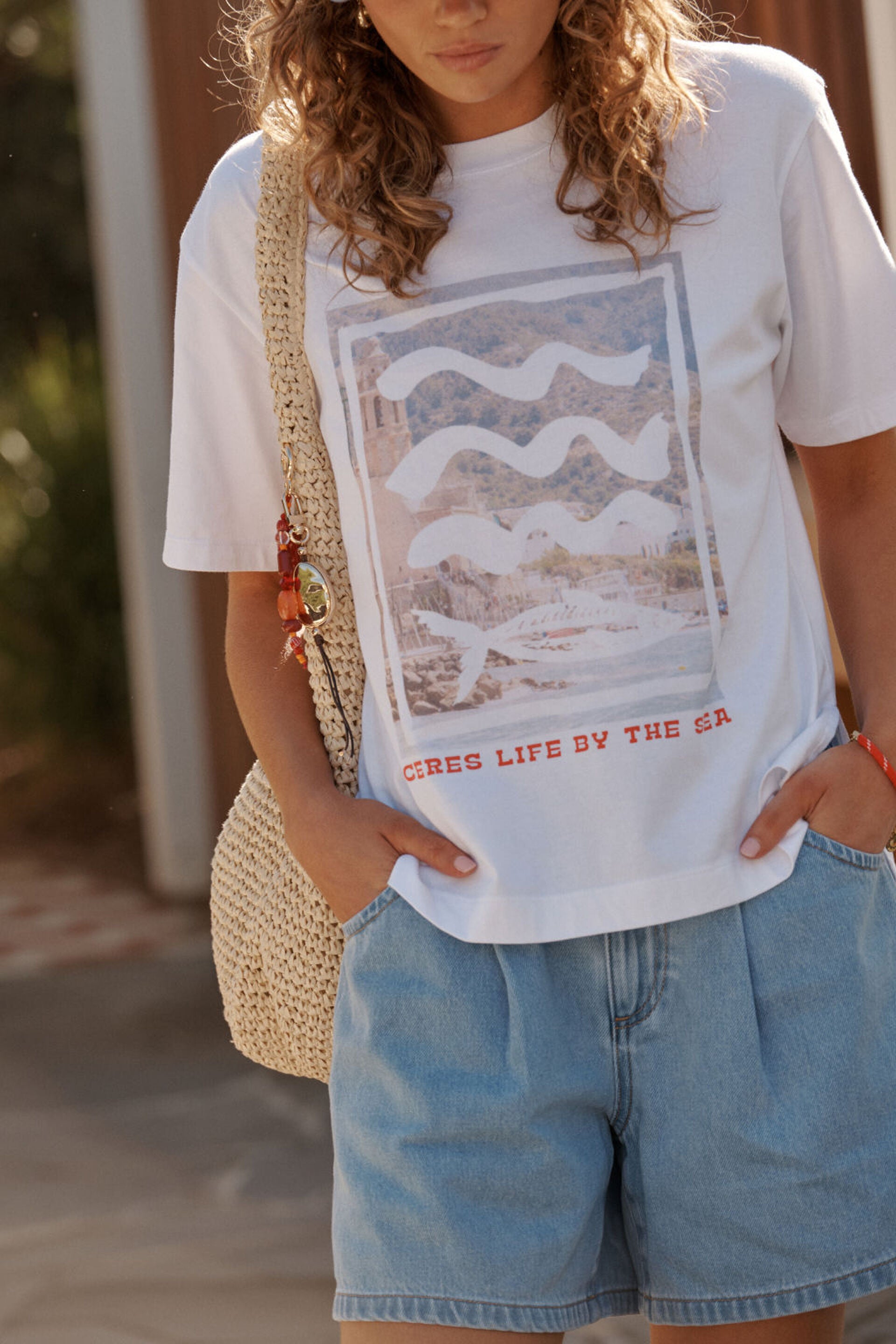 Image of Woman wearing a Ceres Life white t-shirt with graphic design and text, holding a straw bag.