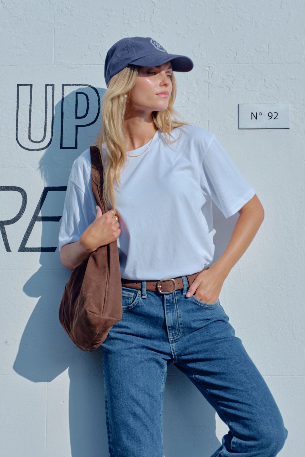 Image of Woman wearing a Ceres Life white t-shirt, blue jeans, and a blue cap against a light gray wall.
