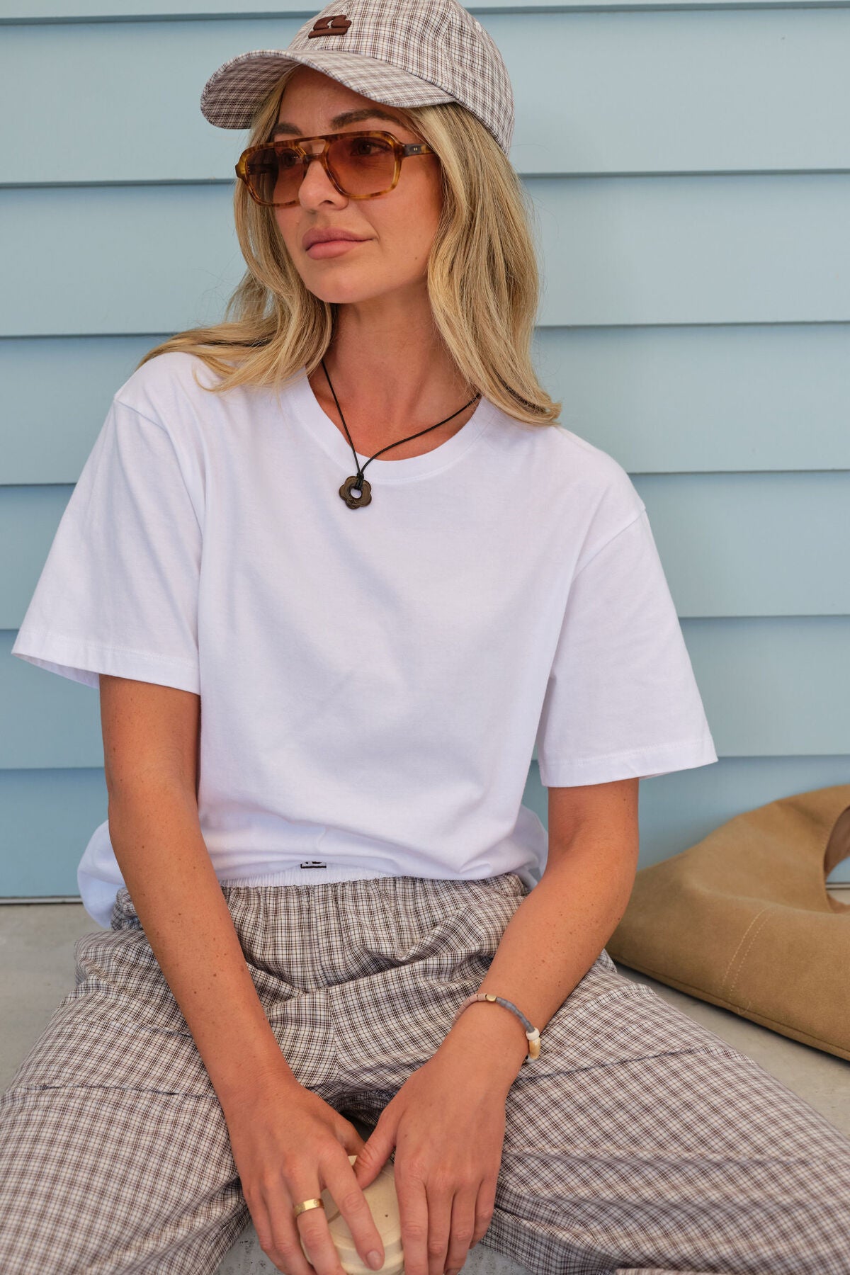 Image of Woman wearing a Ceres Life white t-shirt, plaid pants, and sunglasses sitting against a light blue wall.