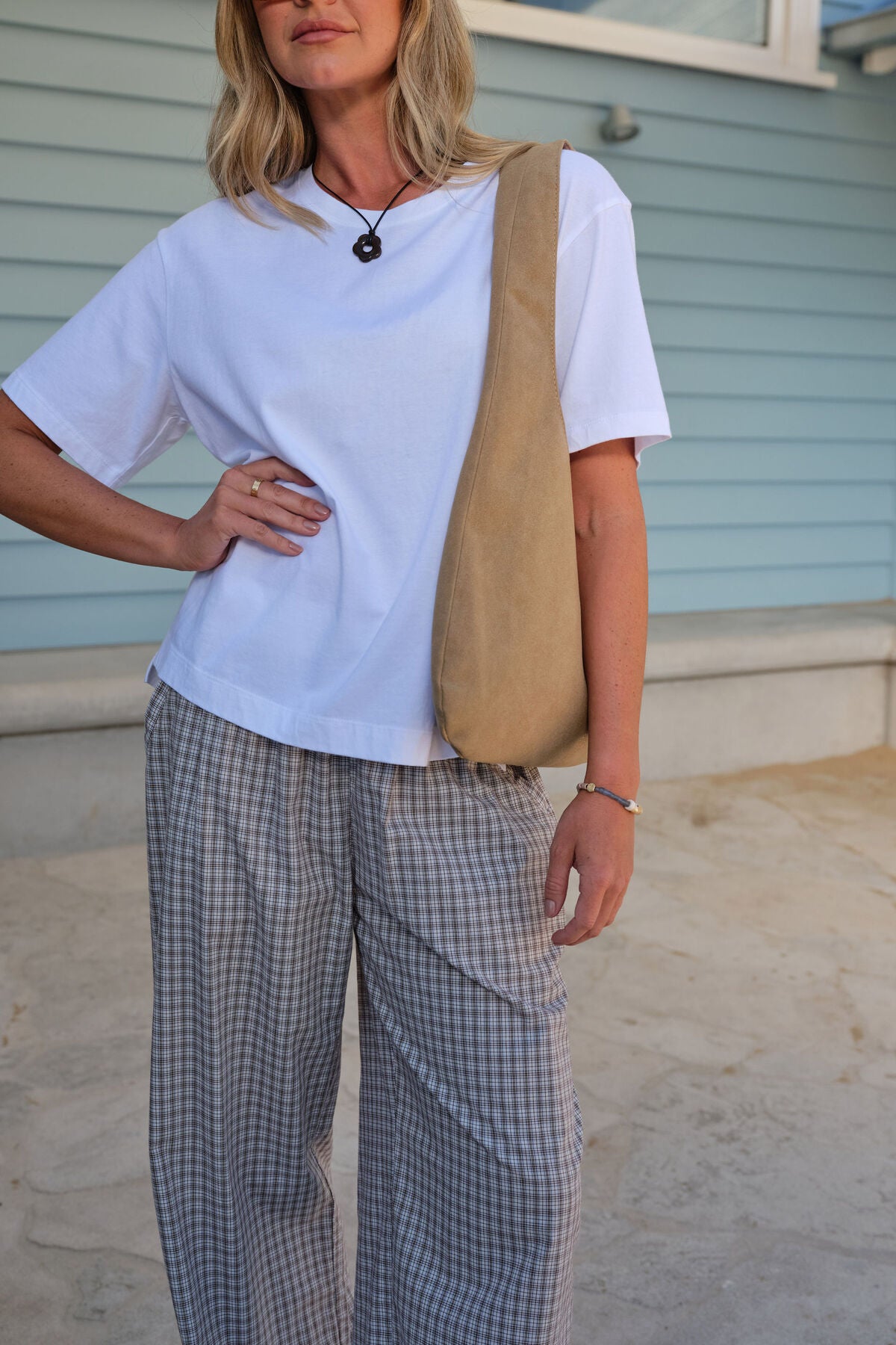 Image of Woman wearing a Ceres Life white t-shirt, beige bag, and checkered pants standing outdoors.