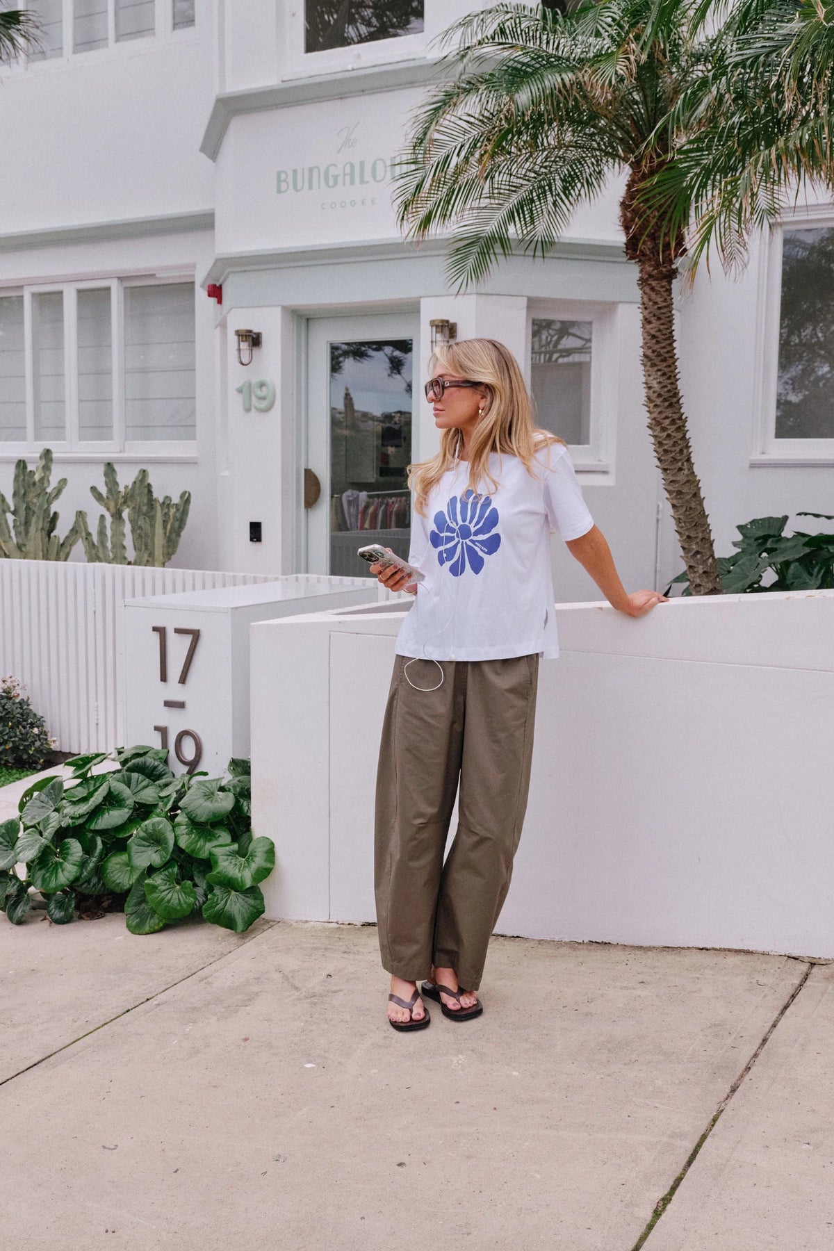 Woman wearing Ceres Life, white tshirt with a feature flower on the front paired with khaki trousers and thongs