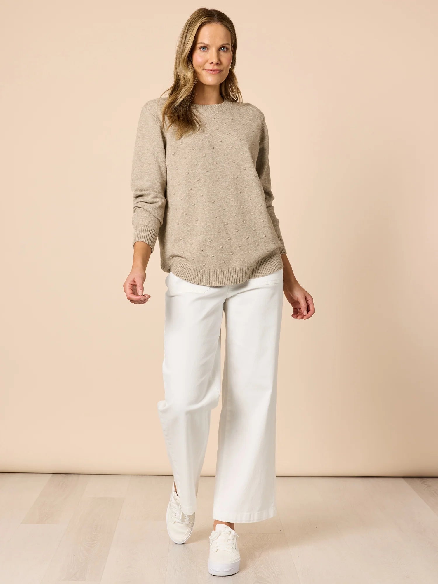 Image of Woman wearing a Clarity beige sweater and white pants against a beige background