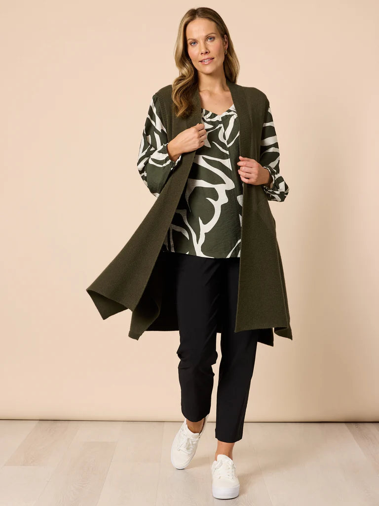 Woman wearing a Clarity long olive green cardigan over a patterned top with black pants and white sneakers on a beige background.